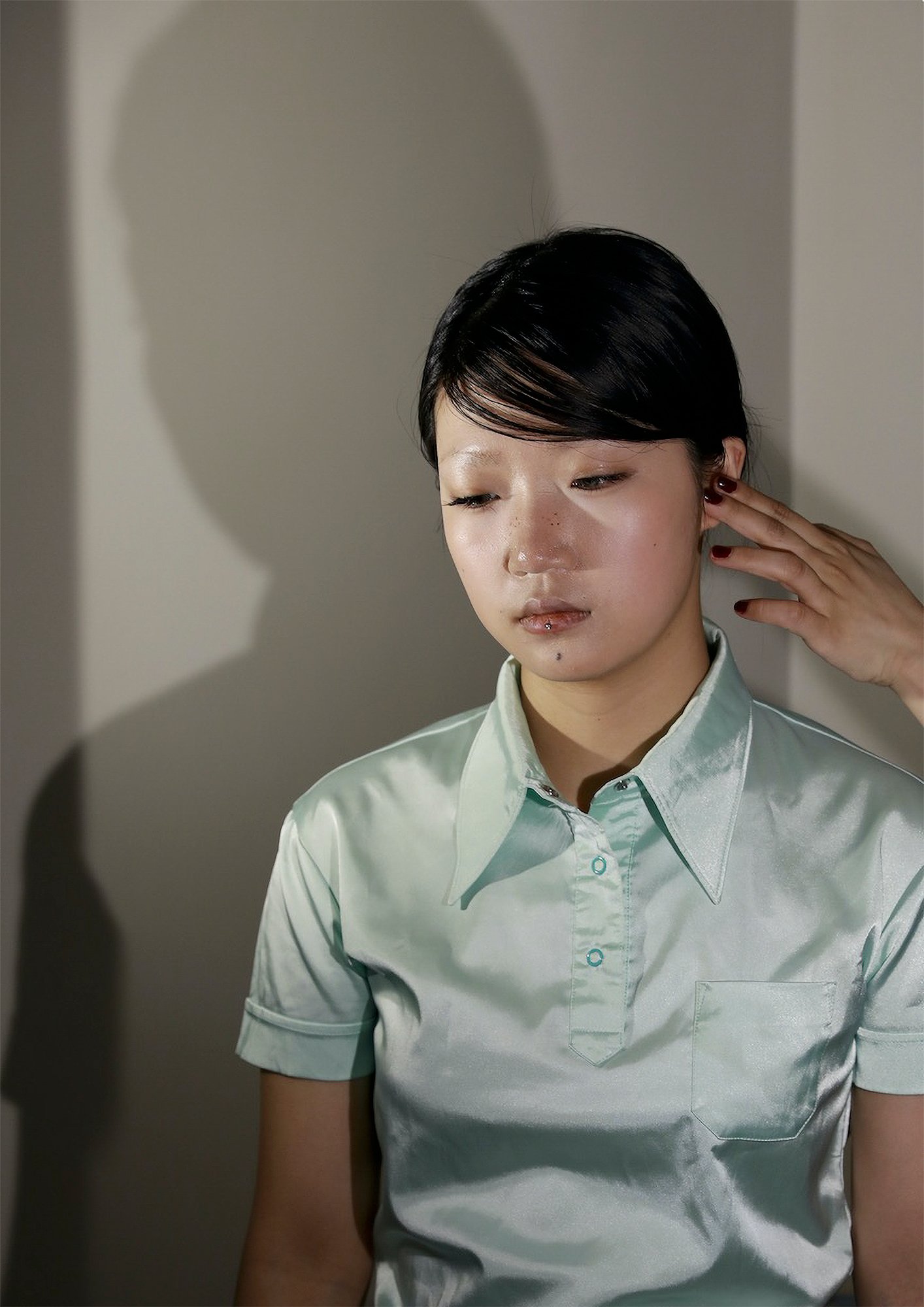 Trust Me by Jinyong Lian, photographed by Jinyong Lian — A woman in a mint-green short-sleeve shirt leans against a white wall, eyes closed, fingers touching her temple, a shadow twin beside her.