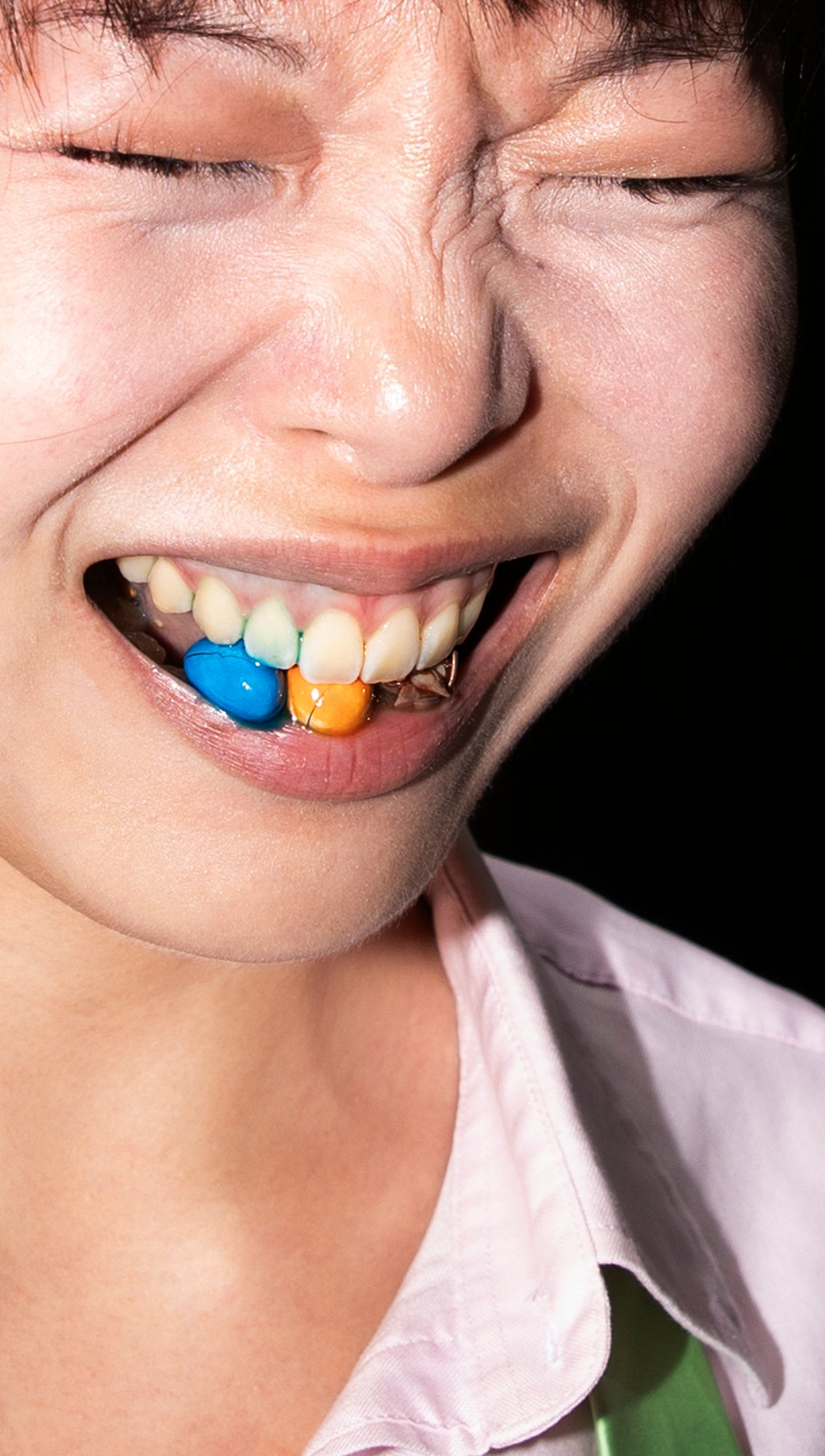 Trust Me by Jinyong Lian, photographed by Jinyong Lian — Close crop of a face grinning wide, coloured candy studs pressed into the gum-line like a grille, black background.