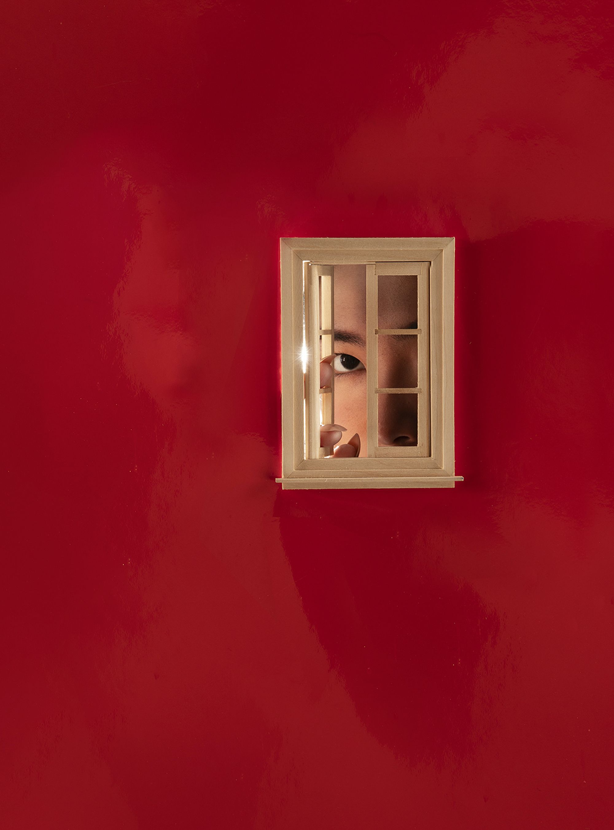 Trust Me by Jinyong Lian, photographed by Jinyong Lian — A face peers through the panes of a tiny dollhouse window set into a deep red lacquered wall, one eye and part of a hand visible.