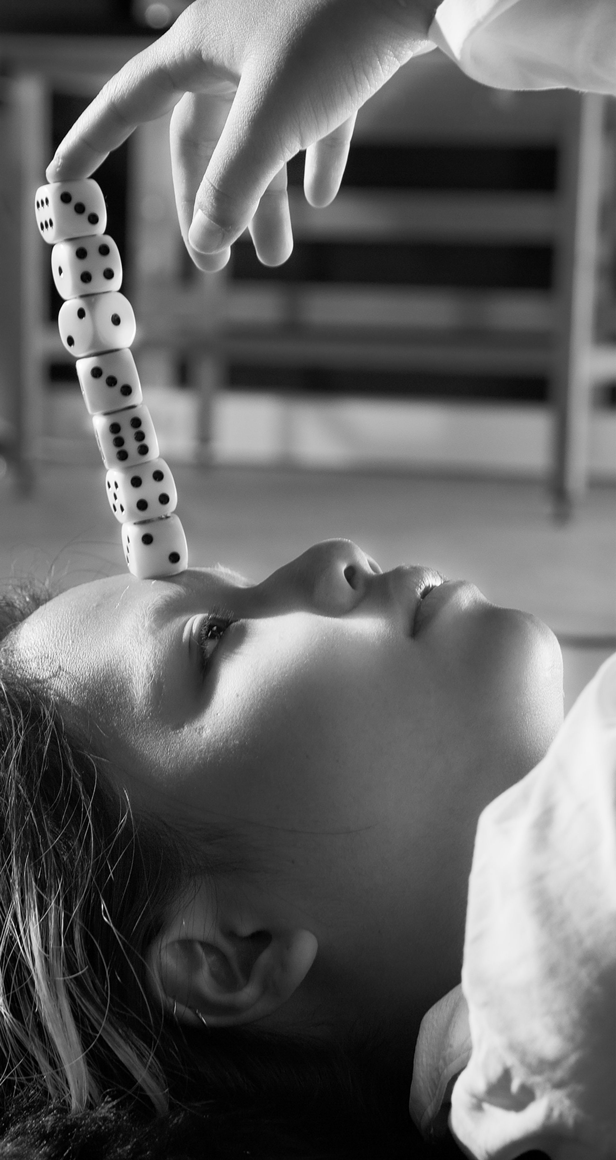 Trust Me by Jinyong Lian, photographed by Jinyong Lian — Black-and-white: a face tipped back, eyes closed, a column of dice balanced nose-bridge to forehead while a gloved hand holds them from above.