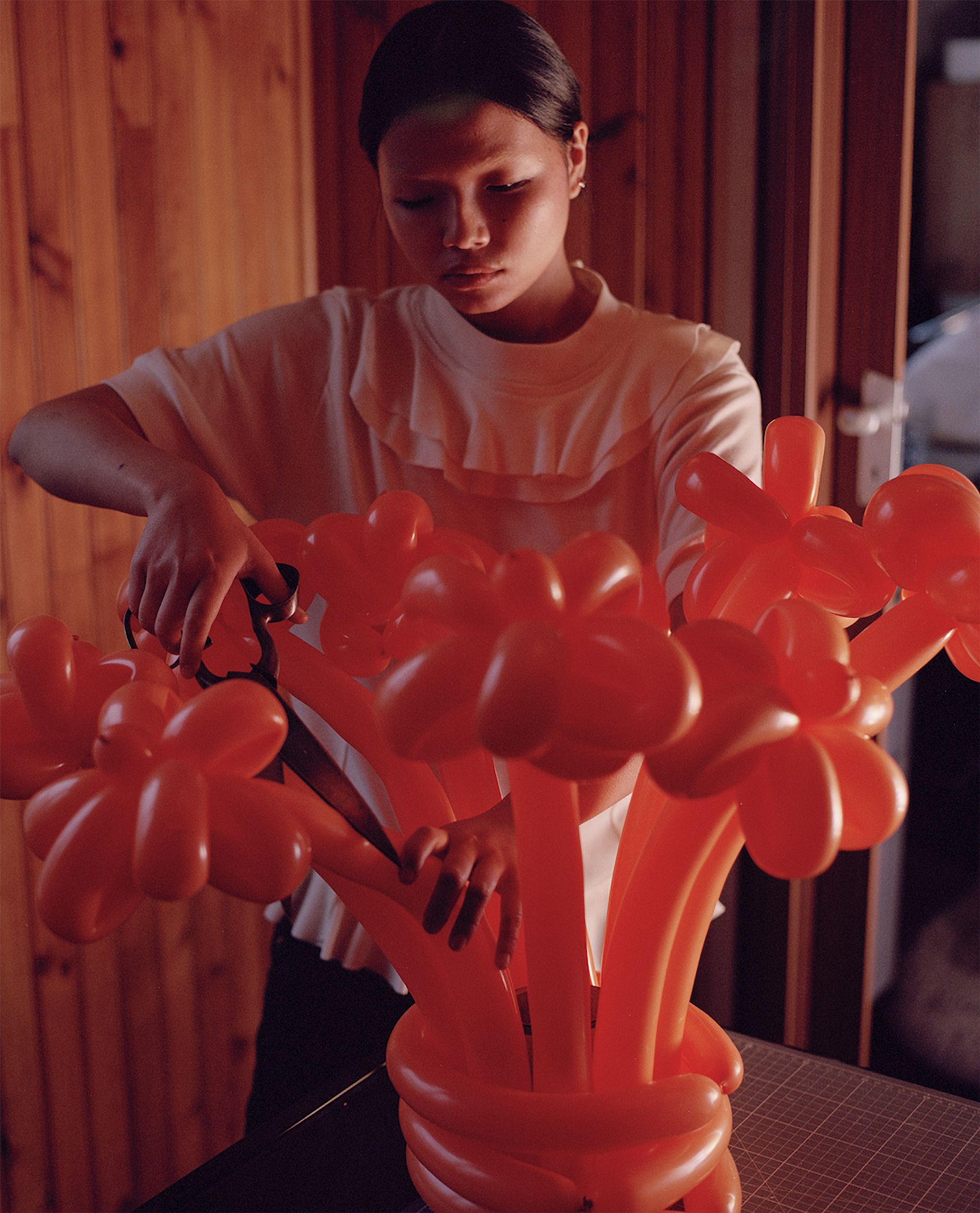 Trust Me by Jinyong Lian, photographed by Jinyong Lian — A woman in a white top works scissors through a mass of orange balloon flowers arranged in a vase, wooden panelling behind.