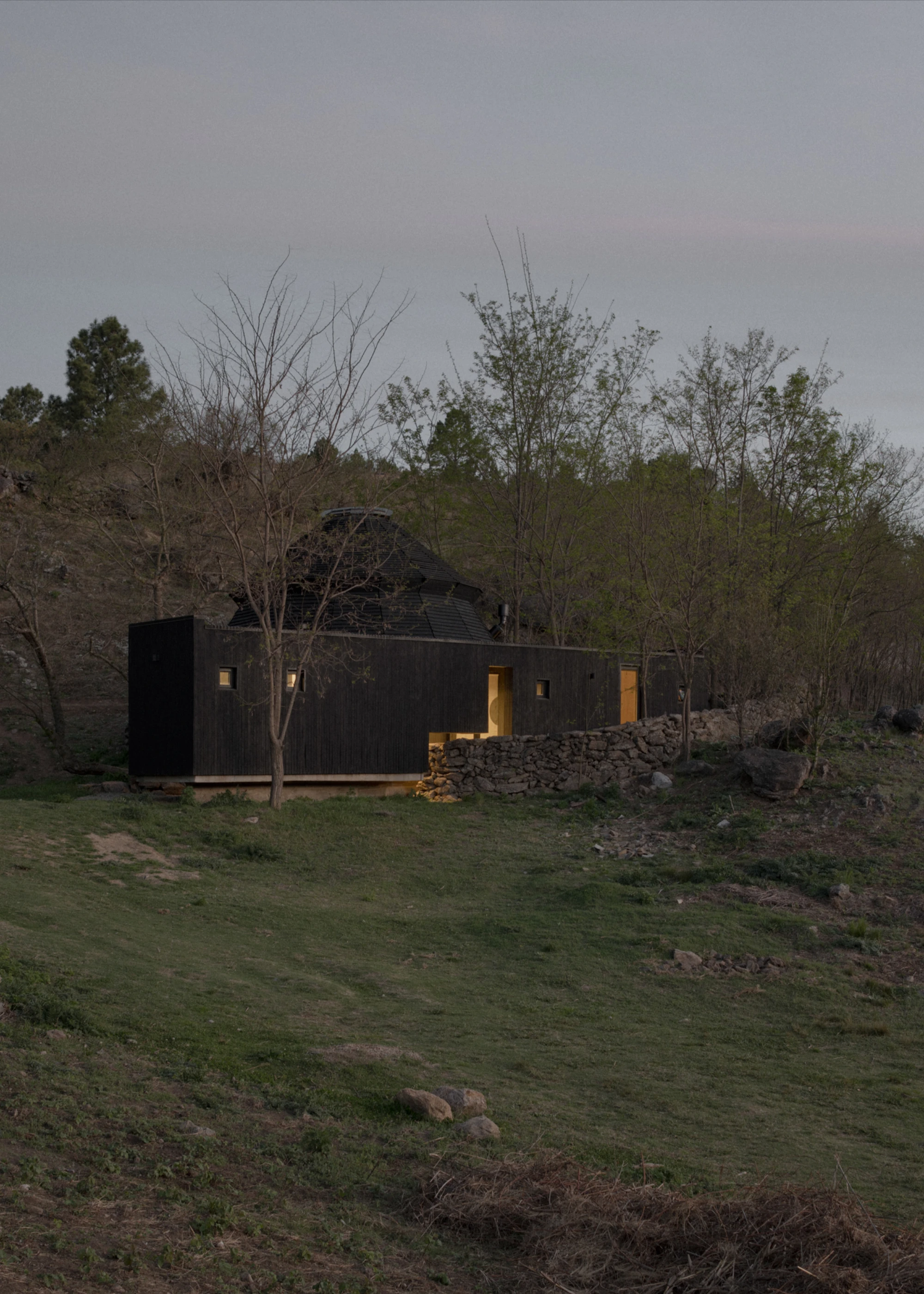 Pavilion El Durazno by Nicolás Oks, photographed by Federico Cairoli — Evening exterior wide shot: service prism windows glow warm amber across the dark hillside, the pavilion dome visible above tree canopy.