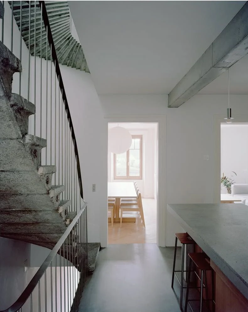 Hameau des Marais by Deschenaux Architectes, photographed by Rory Gardiner — Ground-floor hallway of the lower building: a stone stair with steel balustrade, an exposed concrete beam overhead, the kitchen and dining room visible beyond the door.