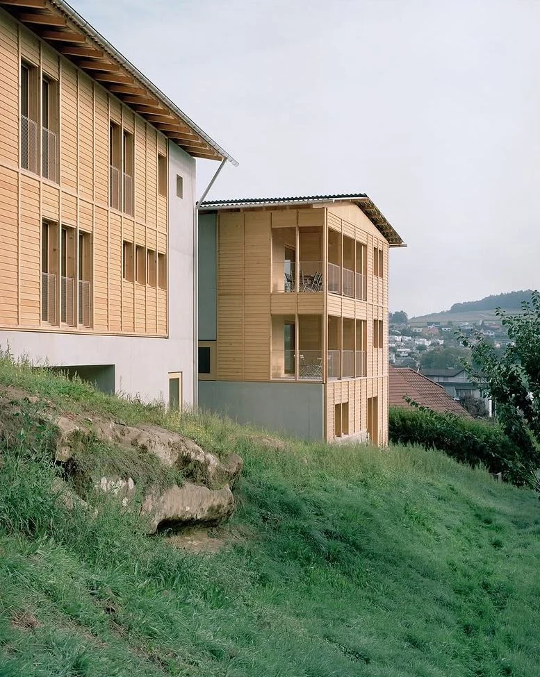 Hameau des Marais by Deschenaux Architectes, photographed by Rory Gardiner — Rear elevation on the slope: the upper glazed loggia open, the larch louvered flanks framing a long view across the Fribourg Mittelland.