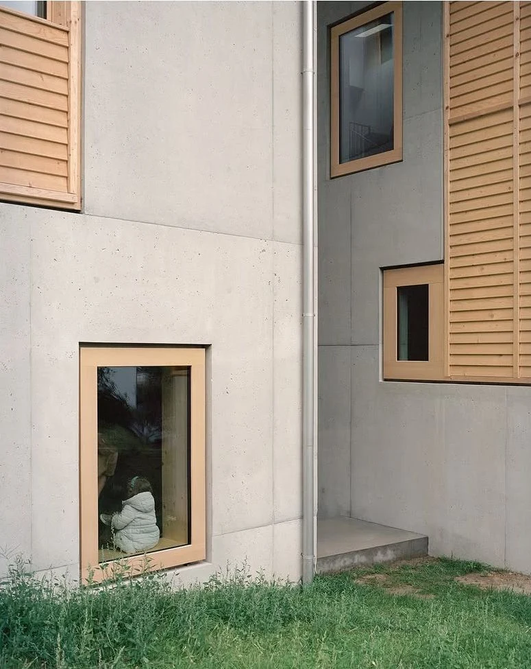 Hameau des Marais by Deschenaux Architectes, photographed by Rory Gardiner — Board-formed concrete wall with a timber-framed window set at low level, a child seated inside silhouetted against the glass, the concrete grain visible across the full panel.