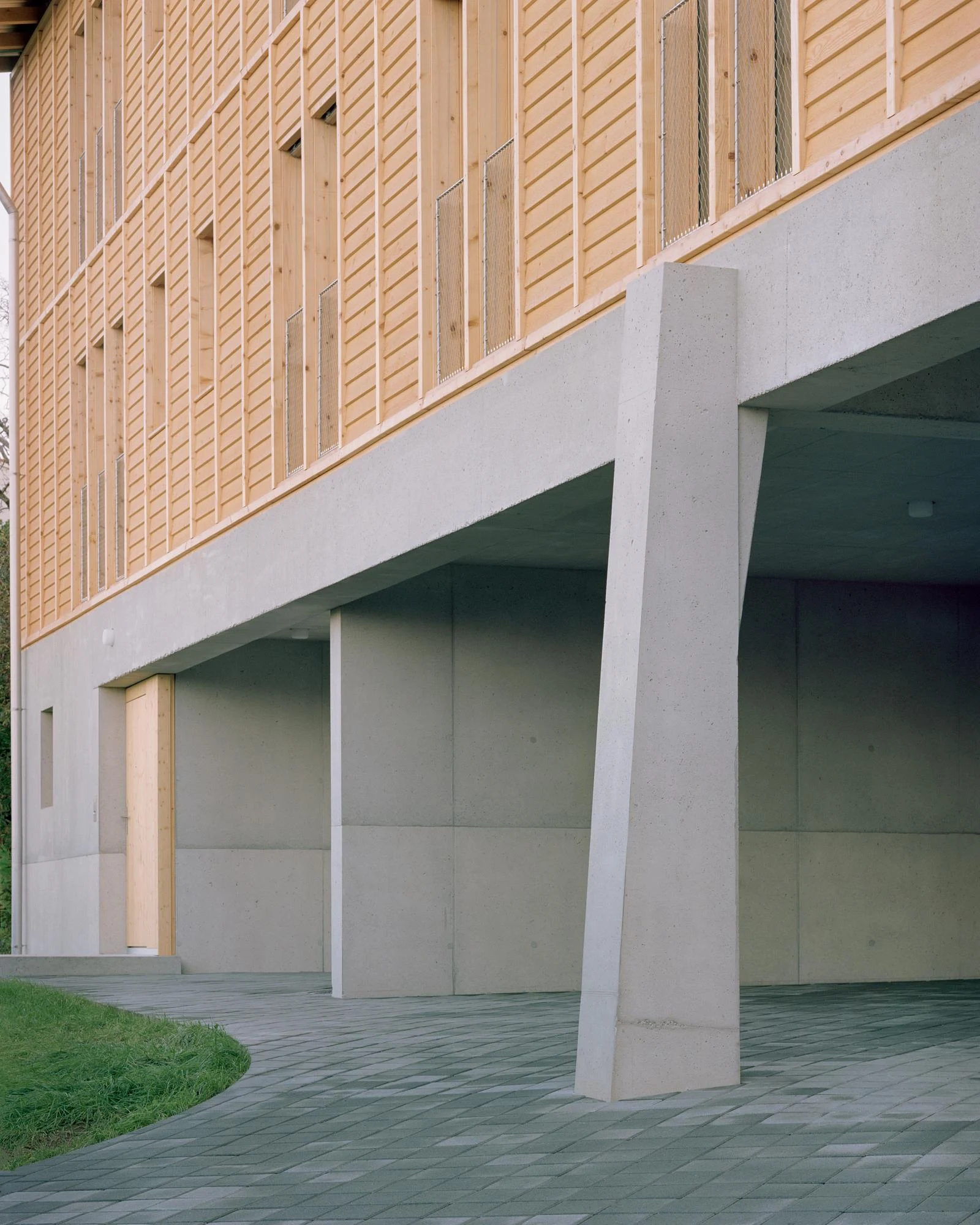 Hameau des Marais by Deschenaux Architectes, photographed by Rory Gardiner — Detail of the concrete pilotis and the larch-louvered skin above: the tapered column and large board-form panel joints read at close range against small-format pavers.
