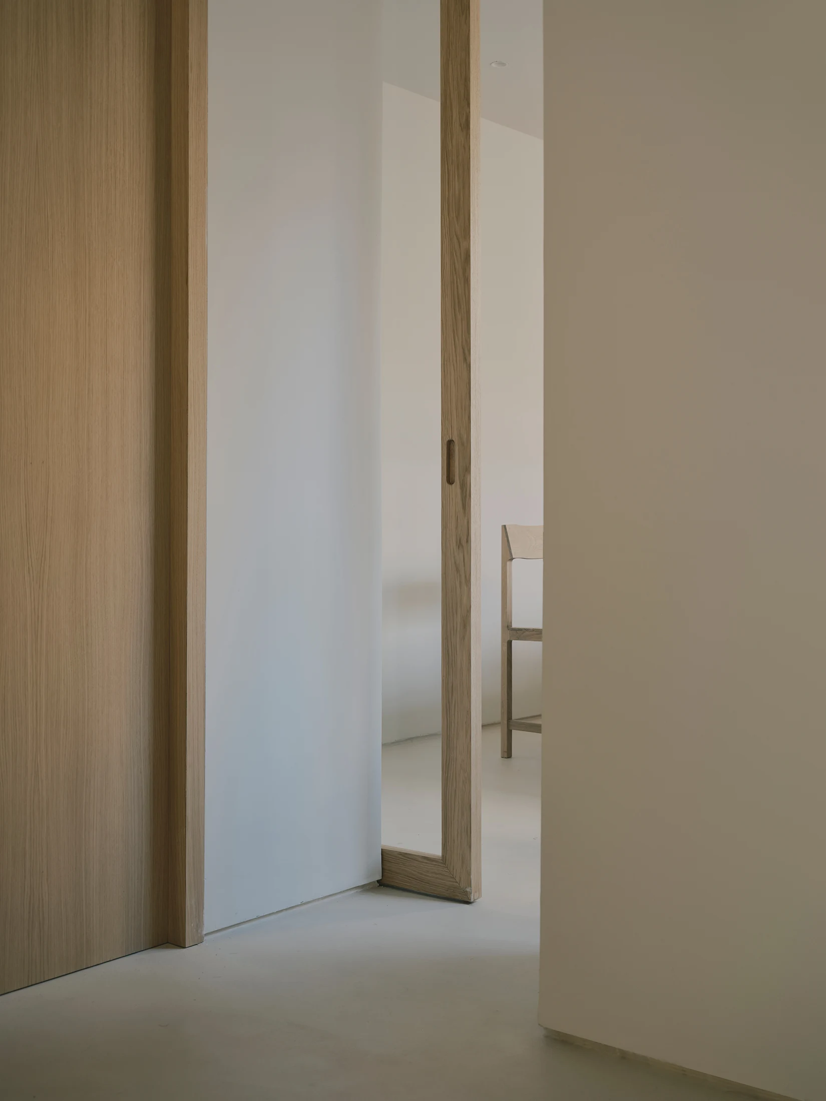Casa St-Maximale by Maximale, photographed by David Zarzoso — Corridor view through two oak-framed doorways in sequence, the frames showing consistent straight grain, microcement floor extending through both openings to a distant room with a narrow oak…