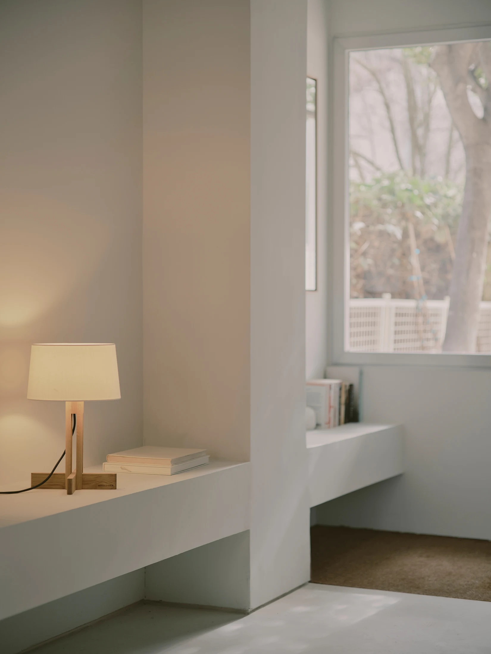 Casa St-Maximale by Maximale, photographed by David Zarzoso — A built-in microcement shelf under a garden-facing window, a wood-framed table lamp on one section and a row of books on a second recessed section, a tree visible through the glazing beyond.