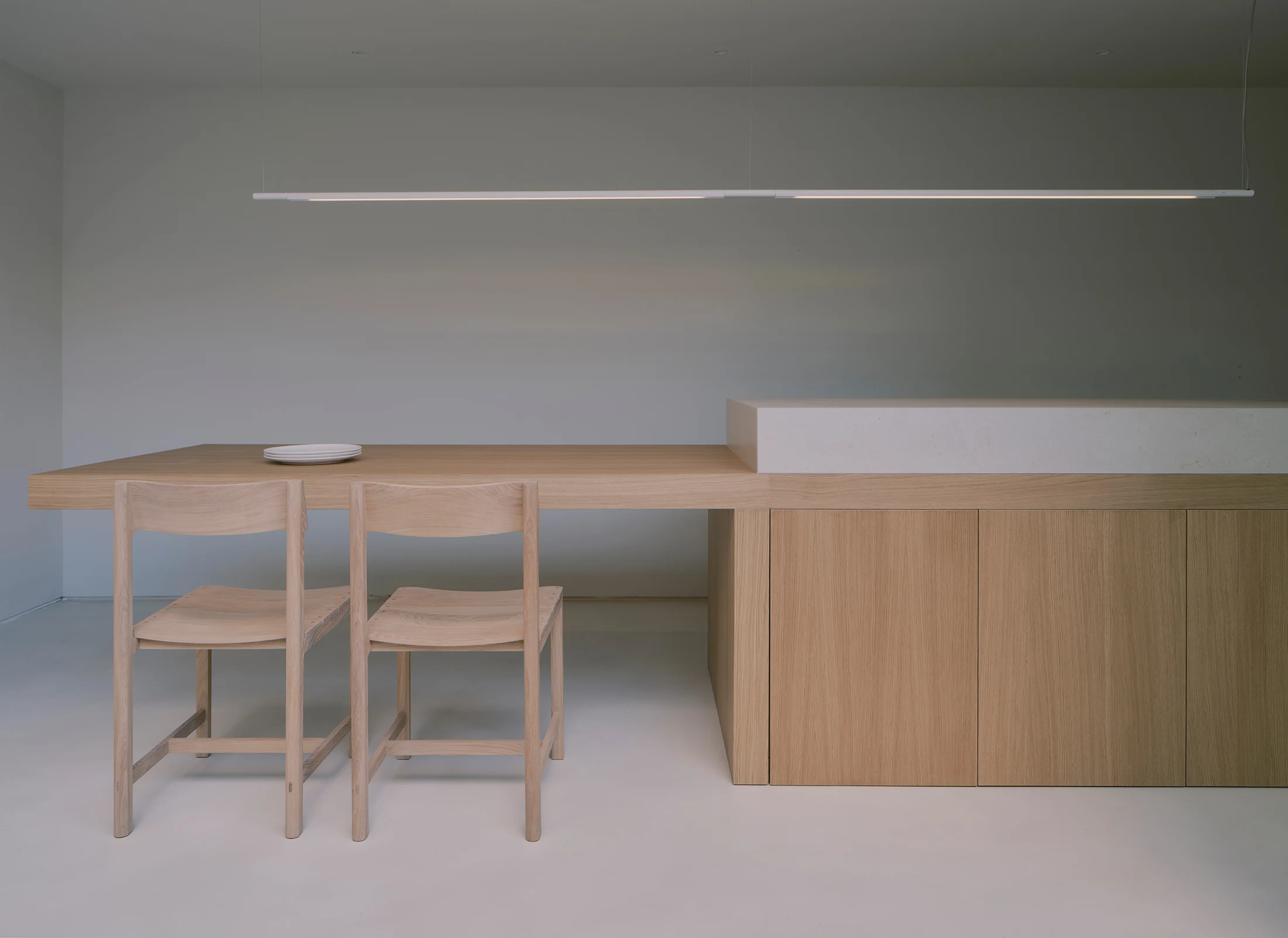 Casa St-Maximale by Maximale, photographed by David Zarzoso — Kitchen-dining viewed from the patio side, the full-height sliding door open, a small olive tree in a raised planter in the foreground, the oak table and kitchen island in the room behind.