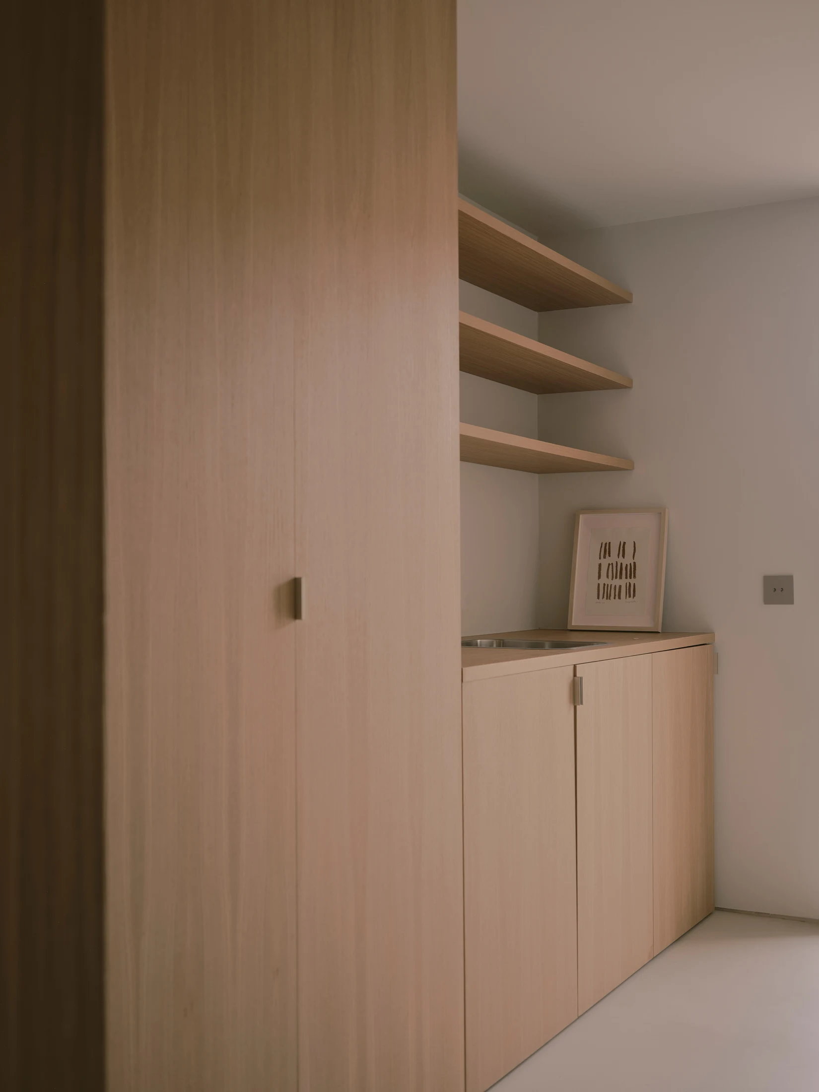 Casa St-Maximale by Maximale, photographed by David Zarzoso — Utility corner with a full-height oak panel door, three floating oak shelves stepping into the corner, and a low oak-fronted base cabinet, microcement floor continuing throughout.