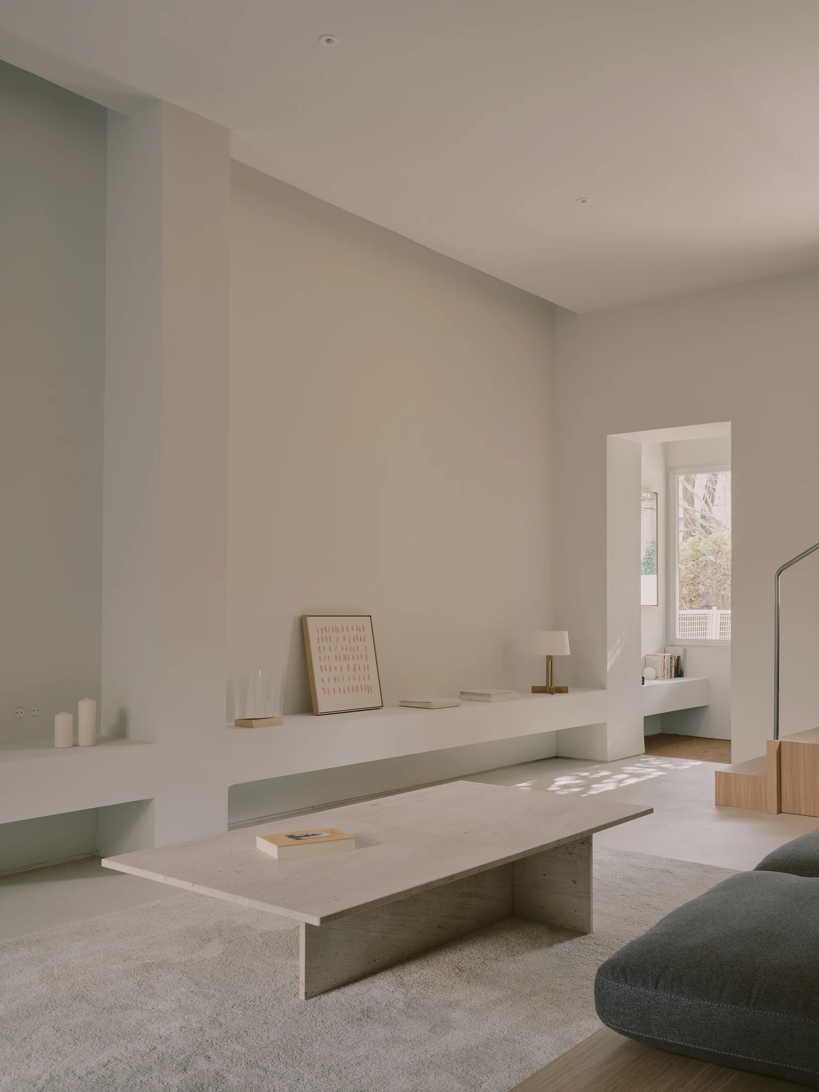 Casa St-Maximale by Maximale, photographed by David Zarzoso — Living room in natural light showing the microcement floor, a travertine-topped coffee table, charcoal sofa, continuous built-in ledge, and an enfiladed view into a second room with garden…