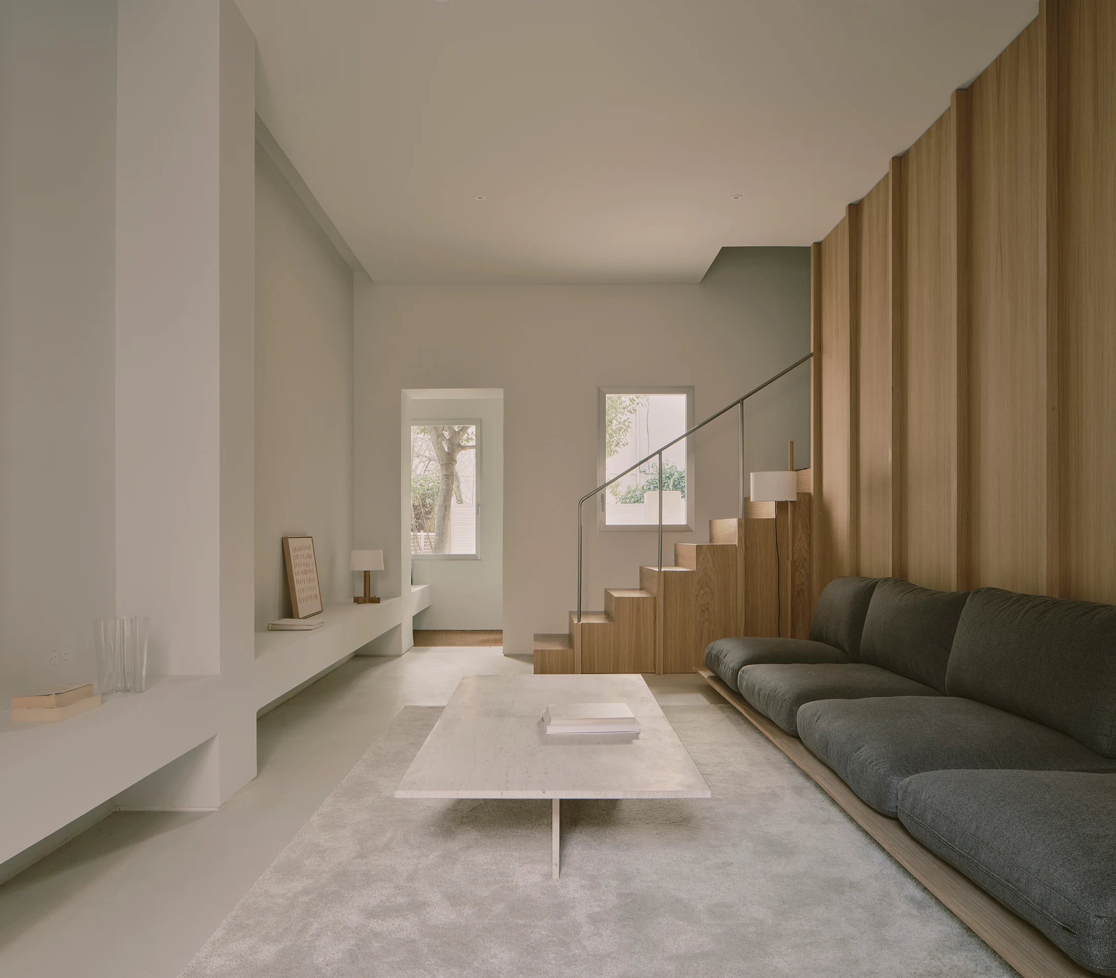 Casa St-Maximale by Maximale, photographed by David Zarzoso — Ground-floor living room with pale microcement floor, a low-profile charcoal sofa, white plaster walls, a solid oak staircase rising against a tall vertical oak-slat panelled wall, two…
