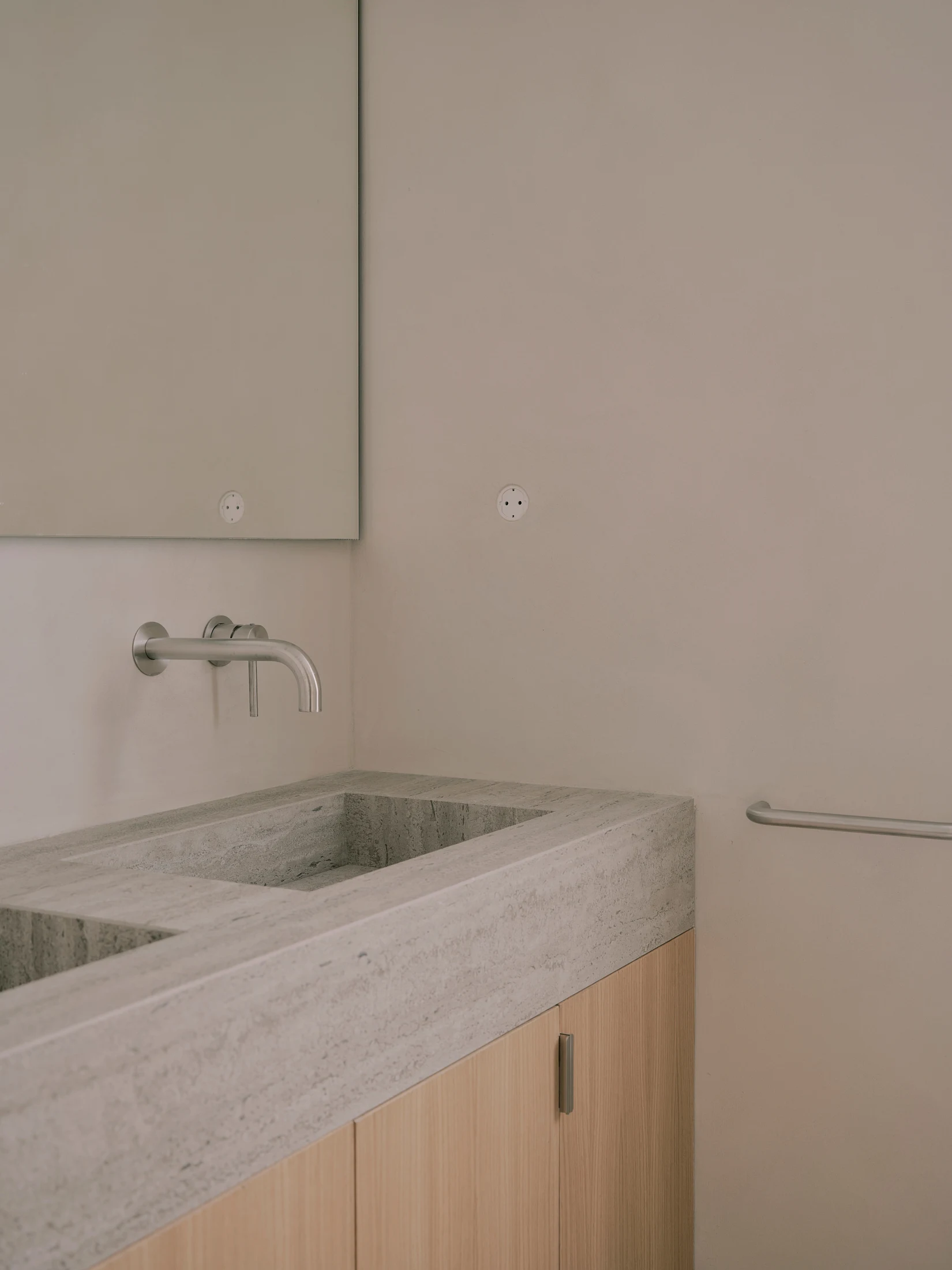 Casa St-Maximale by Maximale, photographed by David Zarzoso — Bathroom vanity detail: a honed grey travertine double-basin top with a wall-mounted brushed steel spout, oak lower cabinet with metal pull, a large mirror above, walls in pale microcement…