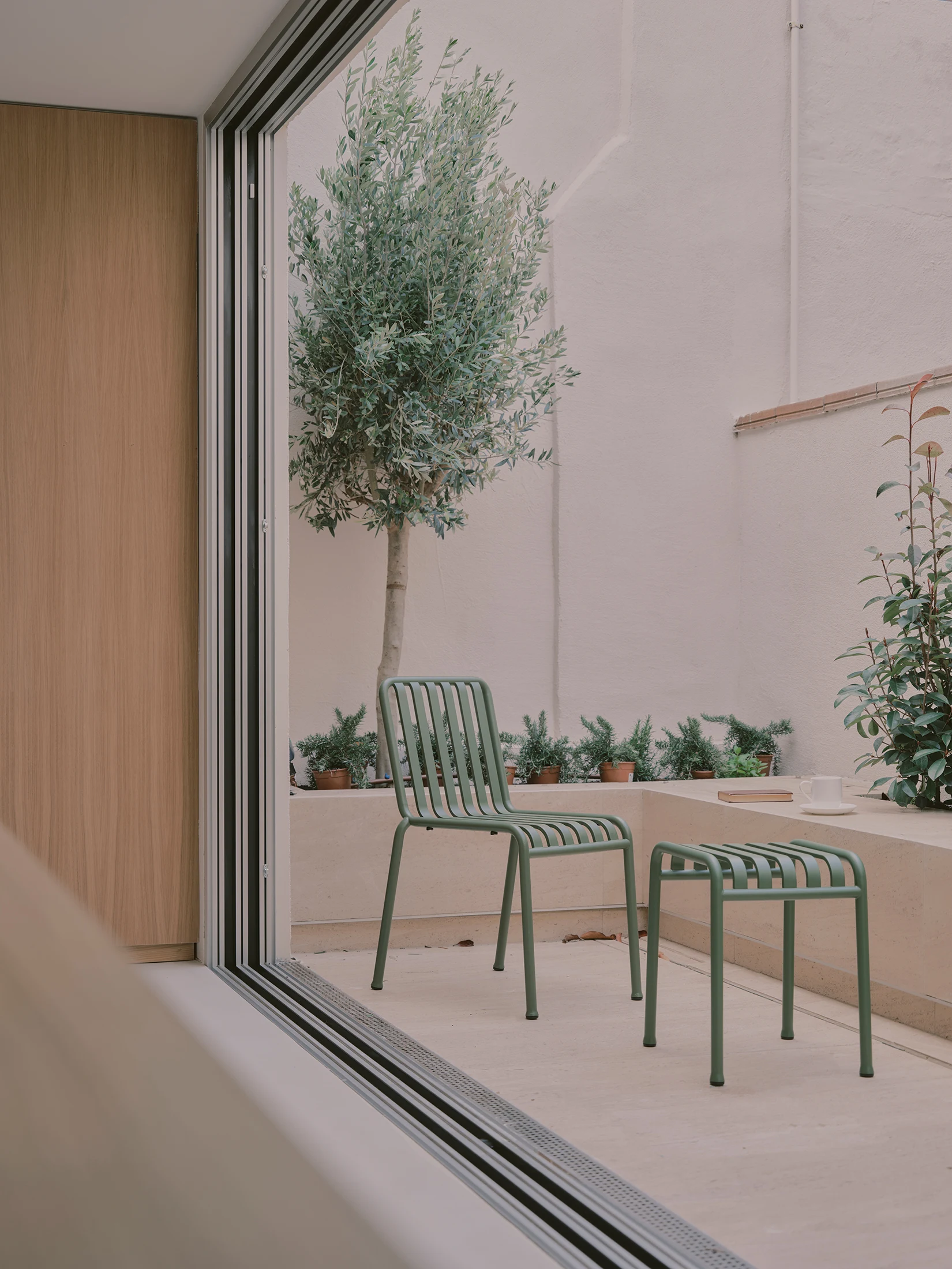 Casa St-Maximale by Maximale, photographed by David Zarzoso — View from kitchen-patio threshold into a ground-floor room beyond, oak slatted wall panel at left edge, sage-green steel tube patio chair and stool in the foreground courtyard.