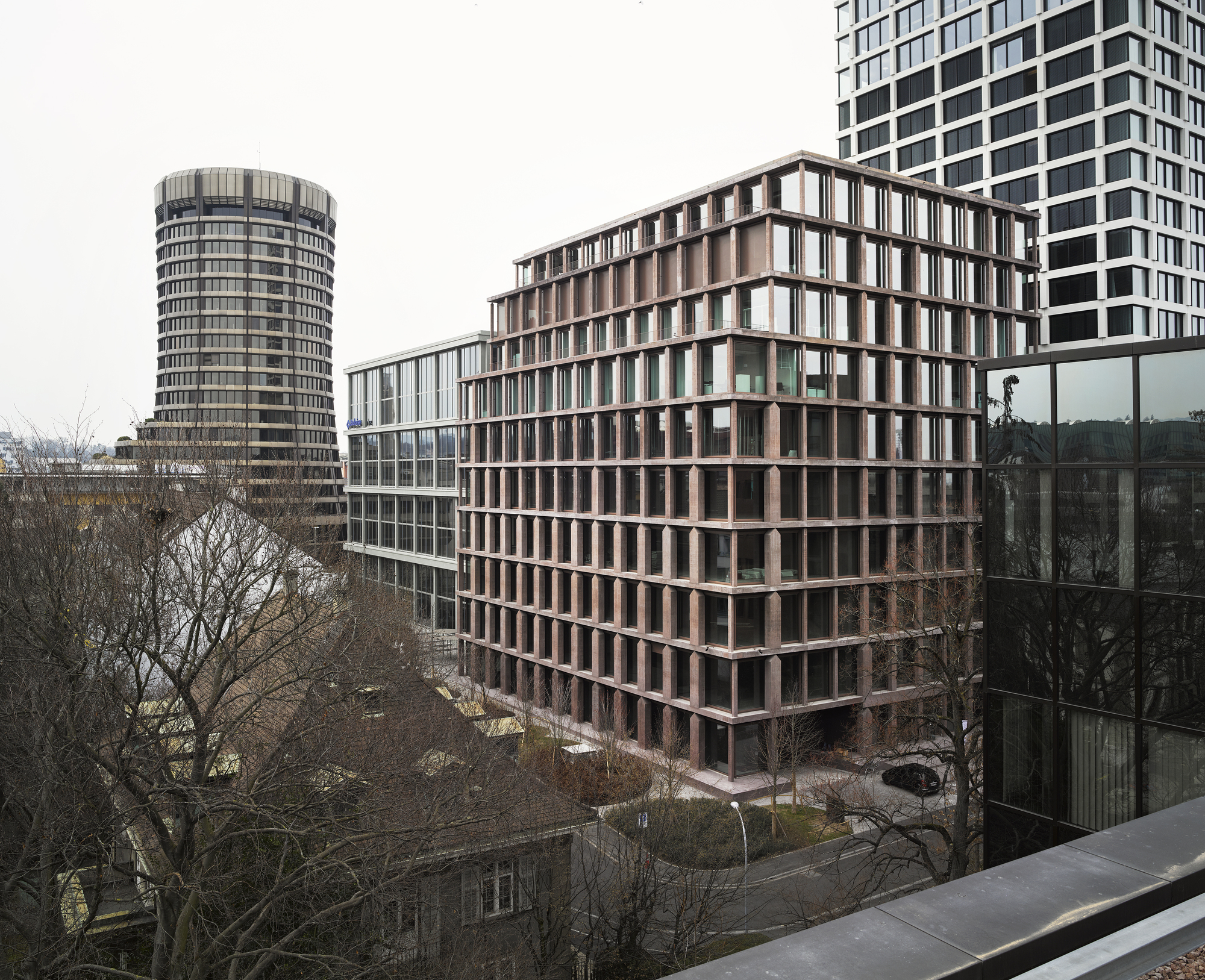 Baloise Insurance Company Office Tower by Valerio Olgiati, photographed by Bas Princen — Brown-pigmented concrete tower rises among Basel's financial district, its house-shaped columns visible against an overcast sky.