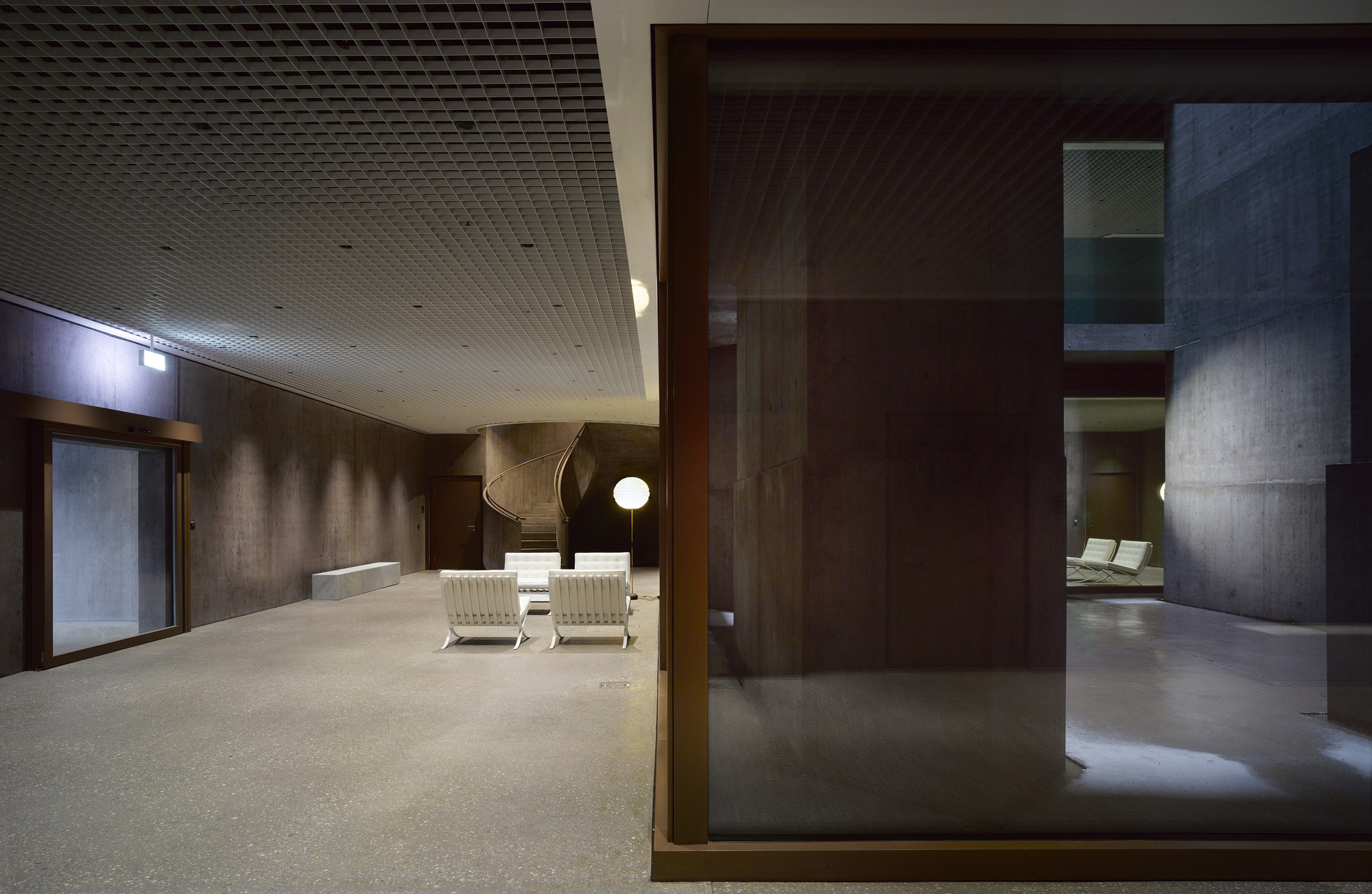 Baloise Insurance Company Office Tower by Valerio Olgiati, photographed by Bas Princen — Lobby space with Barcelona chairs in white leather faces a curved concrete stair, bronze-framed glass enclosures dividing the volume.