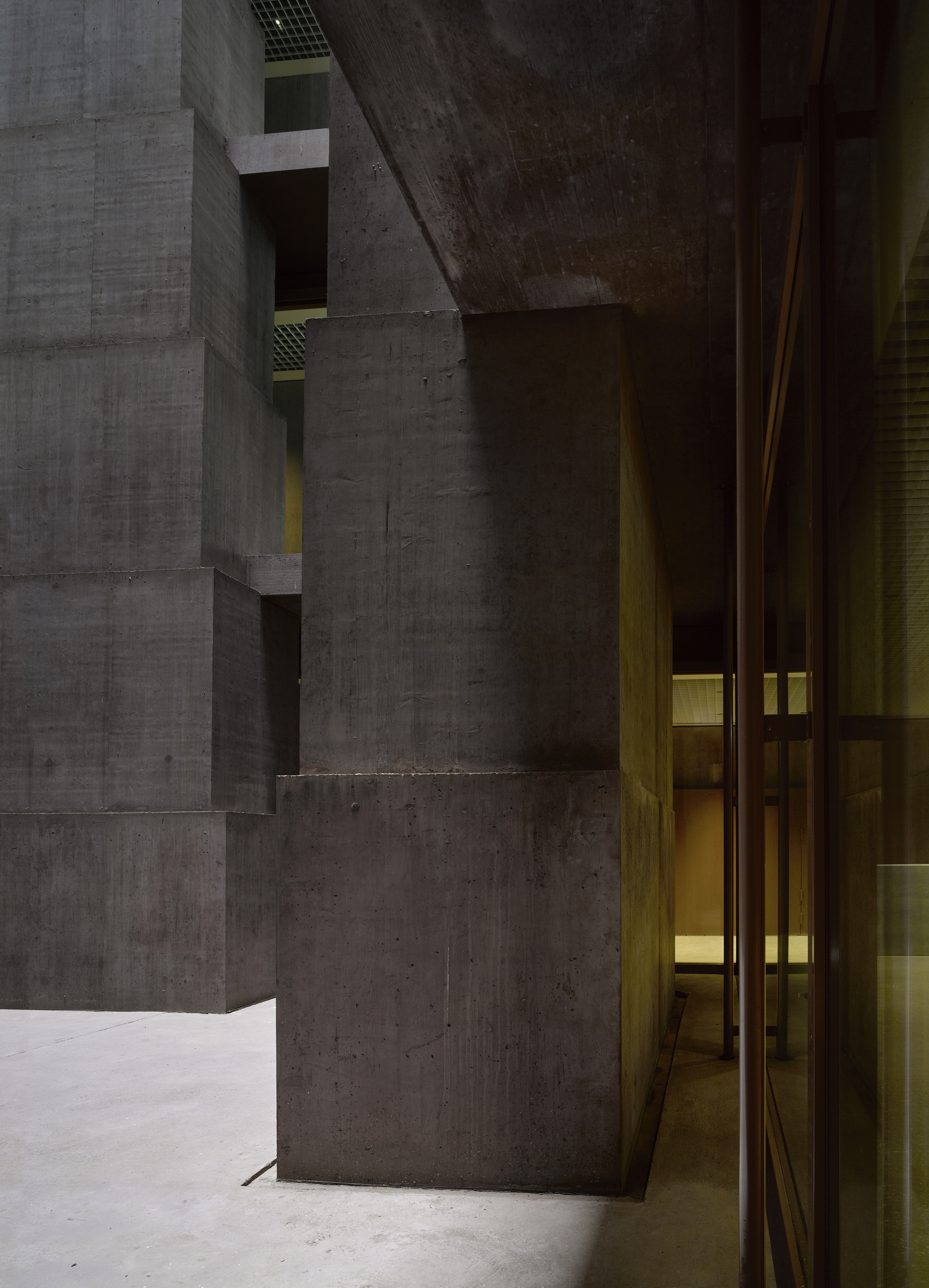Baloise Insurance Company Office Tower by Valerio Olgiati, photographed by Bas Princen — House-shaped column silhouette frames a view toward the glazed lobby, a curved concrete soffit above and warm interior light beyond.