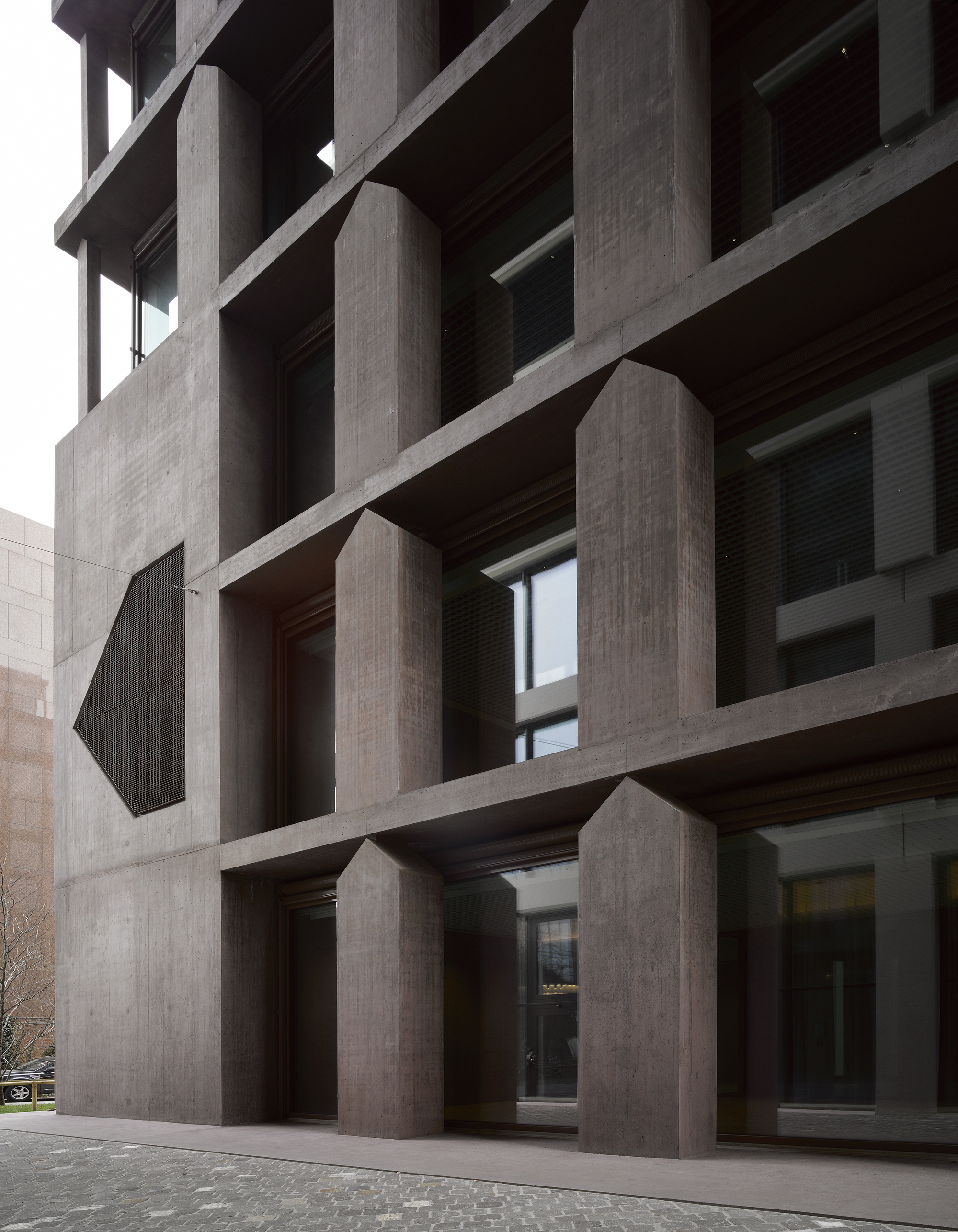 Baloise Insurance Company Office Tower by Valerio Olgiati, photographed by Bas Princen — House-shaped columns in dark brown concrete stand at ground level, their pentagonal profiles creating a primitive silhouette beneath deep horizontal slabs.