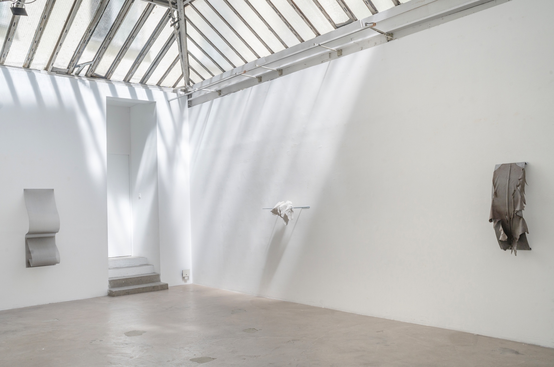 Main de fer, gant de velours by Agurto + Fiedler, photographed by Katherinne Fiedler — Gallery view with skylight trusses visible, plaster wall piece at left, suspended leather with chains at center, folded leather at right.