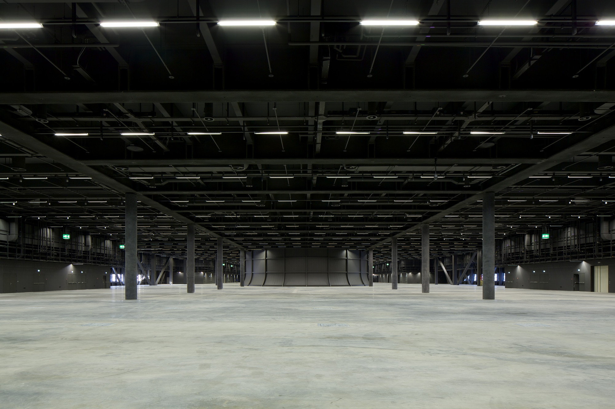 Messe Basel New Hall by Herzog & de Meuron, photographed by Messe Basel — A grid of concrete columns stretches across the empty exhibition hall, linear fluorescent tubes marking rows against the exposed black steel structure.