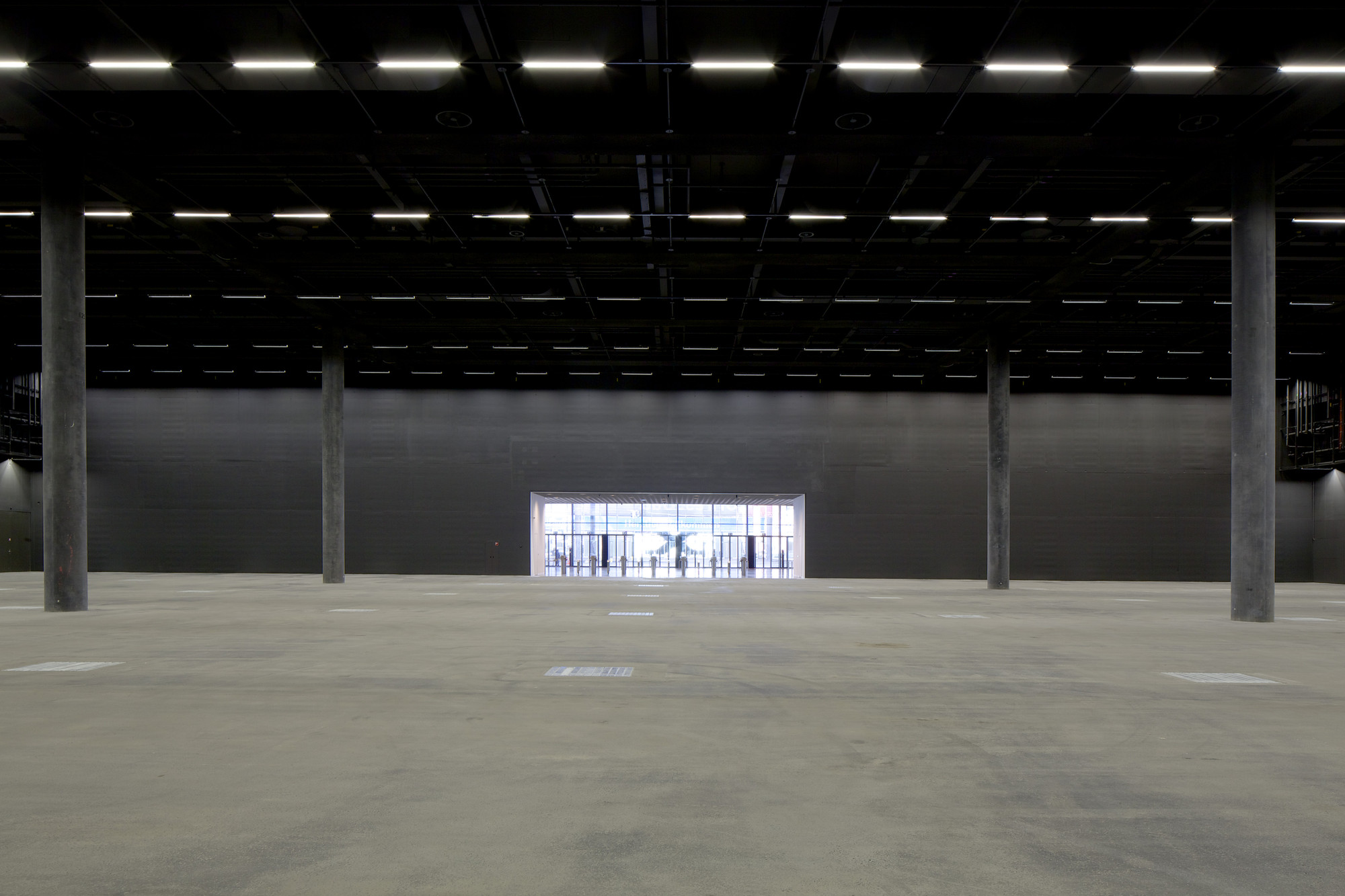 Messe Basel New Hall by Herzog & de Meuron, photographed by Messe Basel — A rectangular opening frames daylight and distant palm trees, concrete columns marching in rhythm across the grey floor toward the exit.