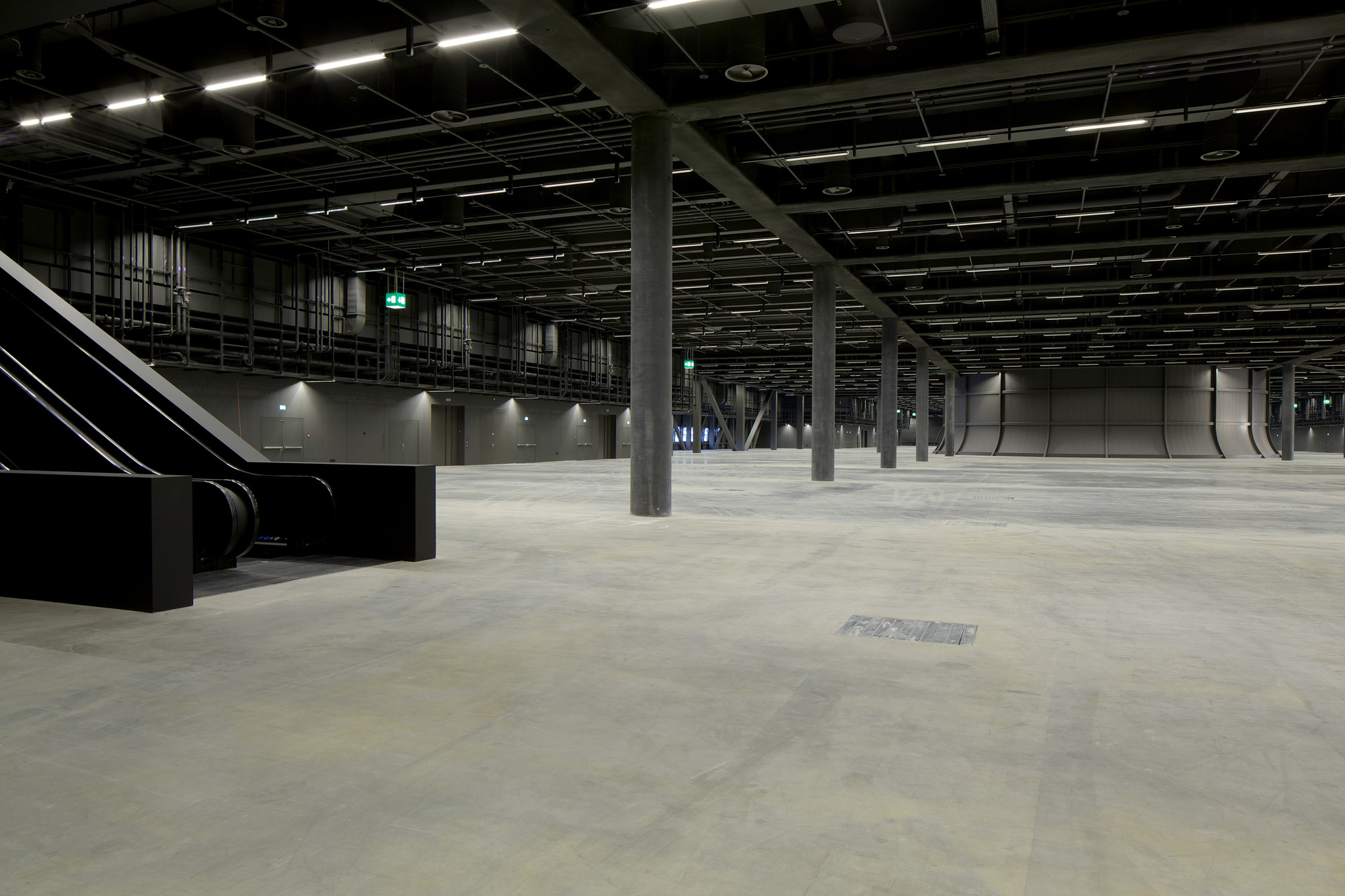 Messe Basel New Hall by Herzog & de Meuron, photographed by Messe Basel — Black-clad escalators angle into the frame, concrete columns receding into the vast hall where curved acoustic panels line the far walls.