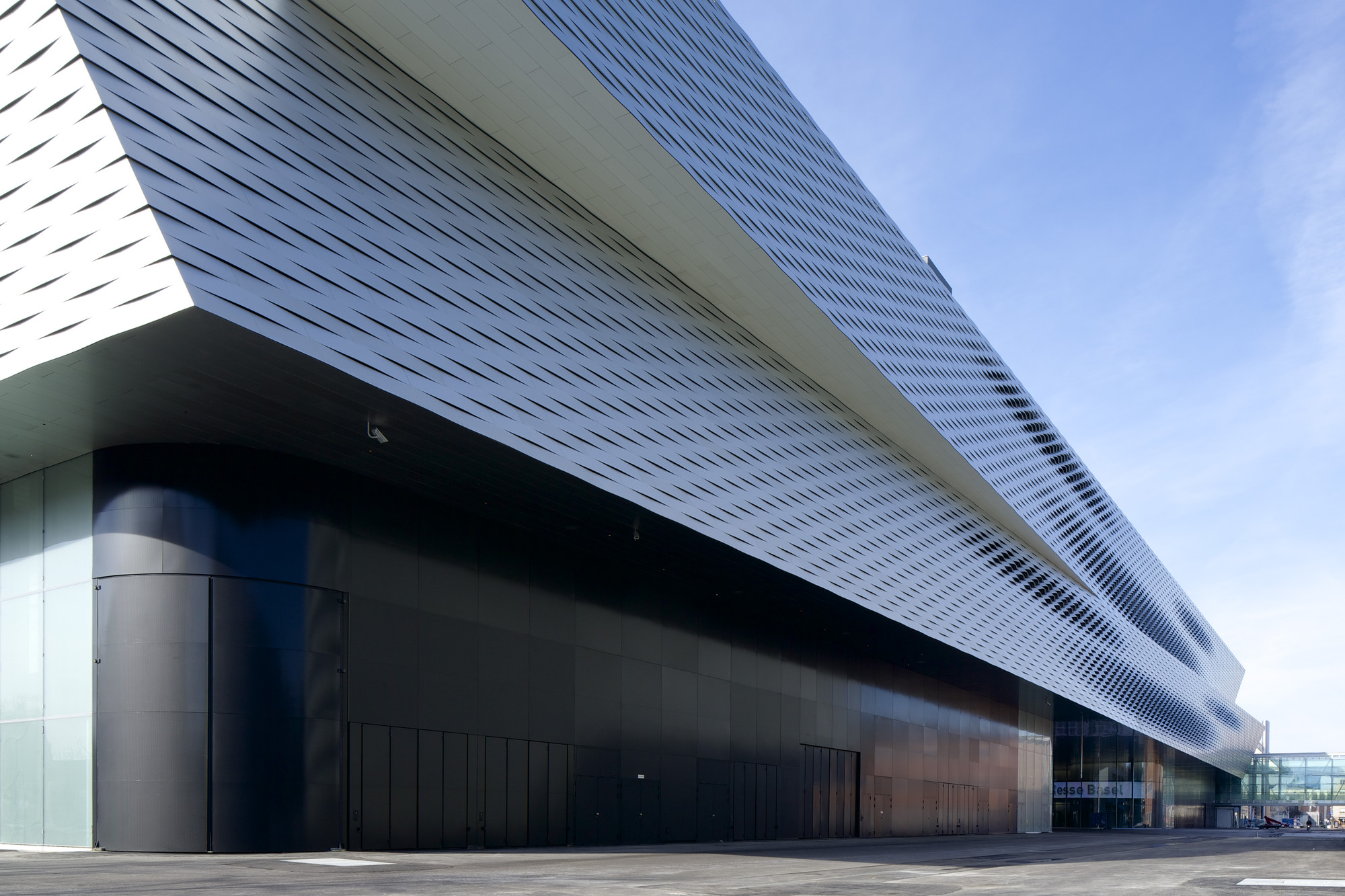 Messe Basel New Hall by Herzog & de Meuron, photographed by Messe Basel — Twisted aluminum bands clad the cantilevered upper volume, their diagonal rhythm catching afternoon light against a dark metal ground floor.