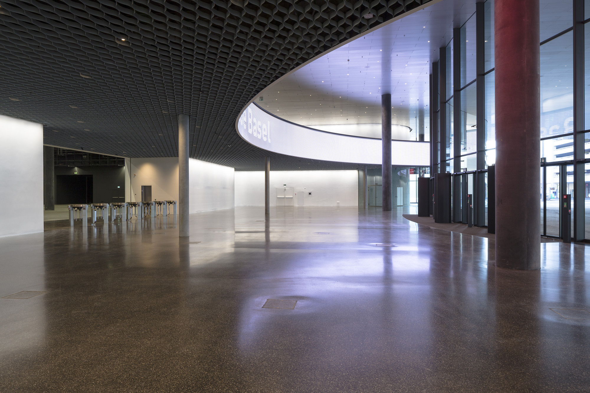Messe Basel New Hall by Herzog & de Meuron, photographed by Messe Basel — The central oculus curves overhead with illuminated Basel signage, polished brown terrazzo floor meeting dark coffered ceiling panels.