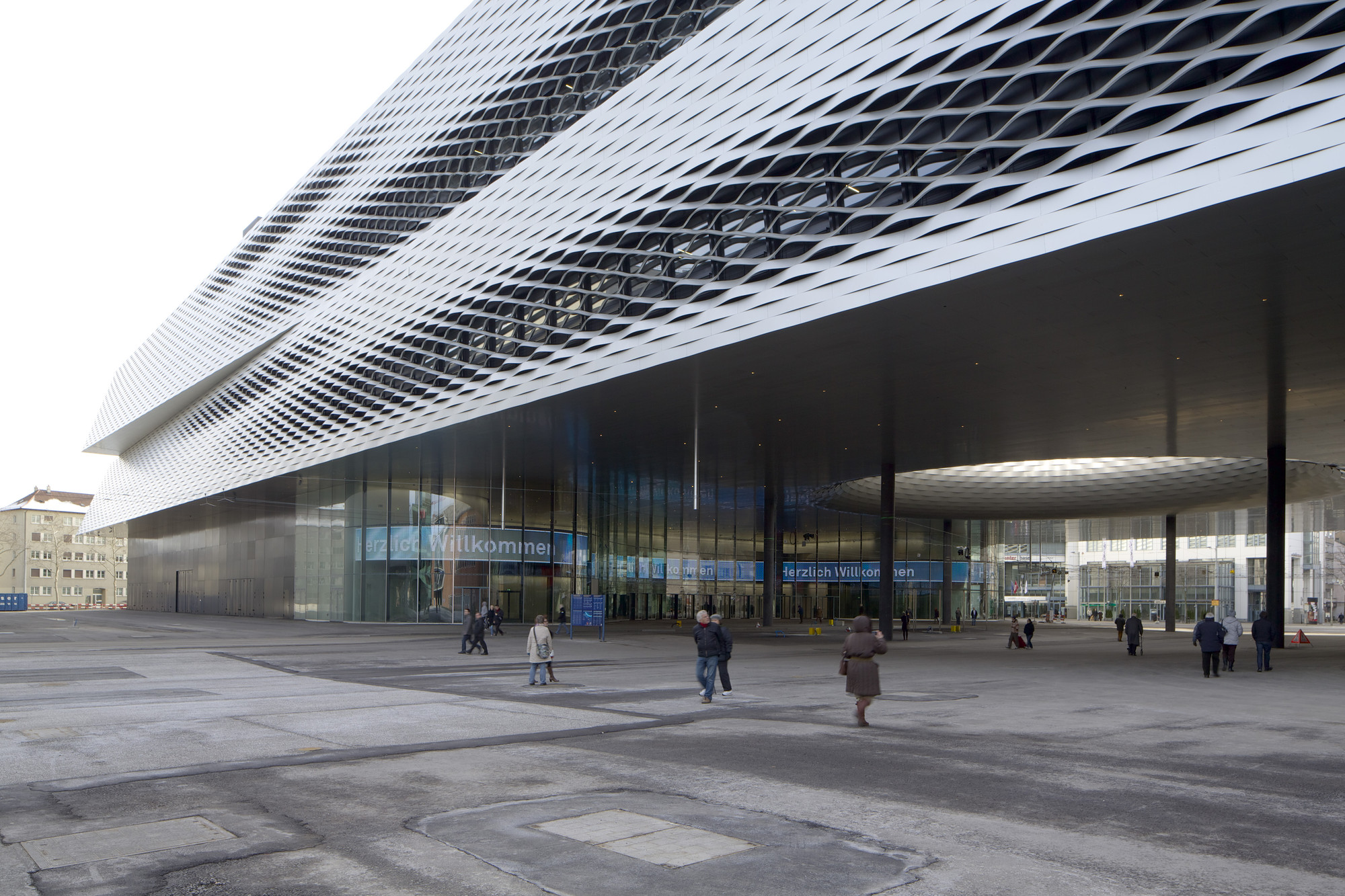 Messe Basel New Hall by Herzog & de Meuron, photographed by Messe Basel — Pedestrians cross the Messeplatz beneath the suspended mass, glass ground floor revealing the covered City Lounge beyond.