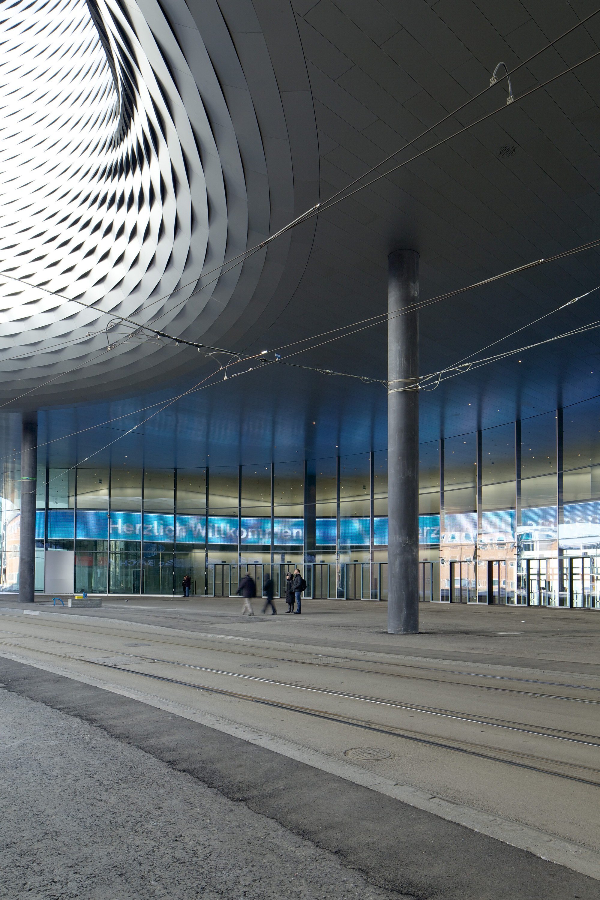 Messe Basel New Hall by Herzog & de Meuron, photographed by Messe Basel — Curved aluminum panels ripple across the City Lounge ceiling, circular concrete columns framing the entrance as tram tracks cut through the paving.
