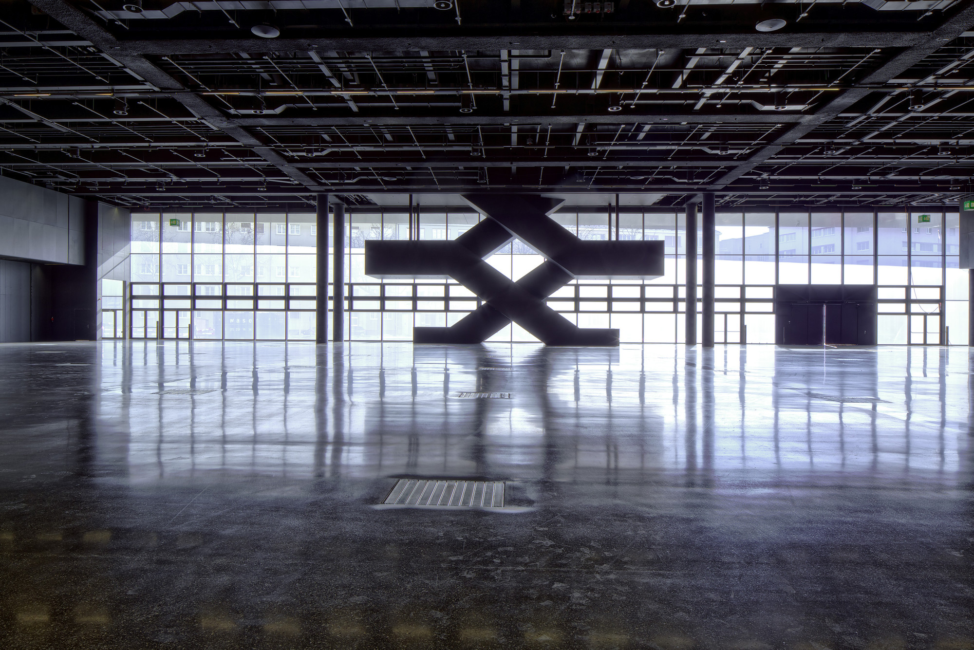 Messe Basel New Hall by Herzog & de Meuron, photographed by Messe Basel — A steel X-brace silhouettes against floor-to-ceiling glazing, polished concrete floor reflecting backlighting from the exhibition hall perimeter.