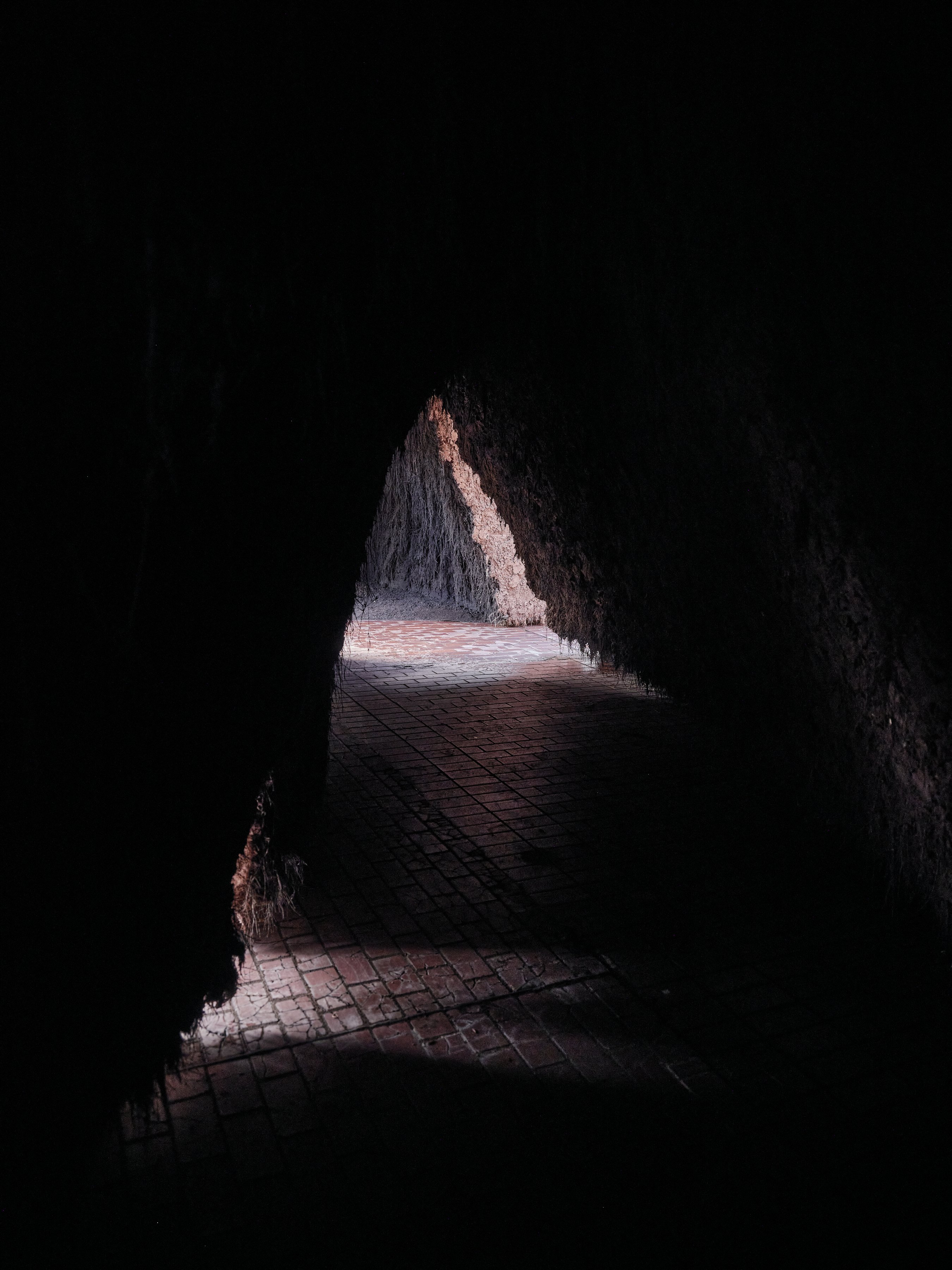 origo by Delcy Morelos, photographed by Thomas Adank — Interior darkness with a triangle of light from a distant opening falling on brick pavement, earth walls absorbing all else.