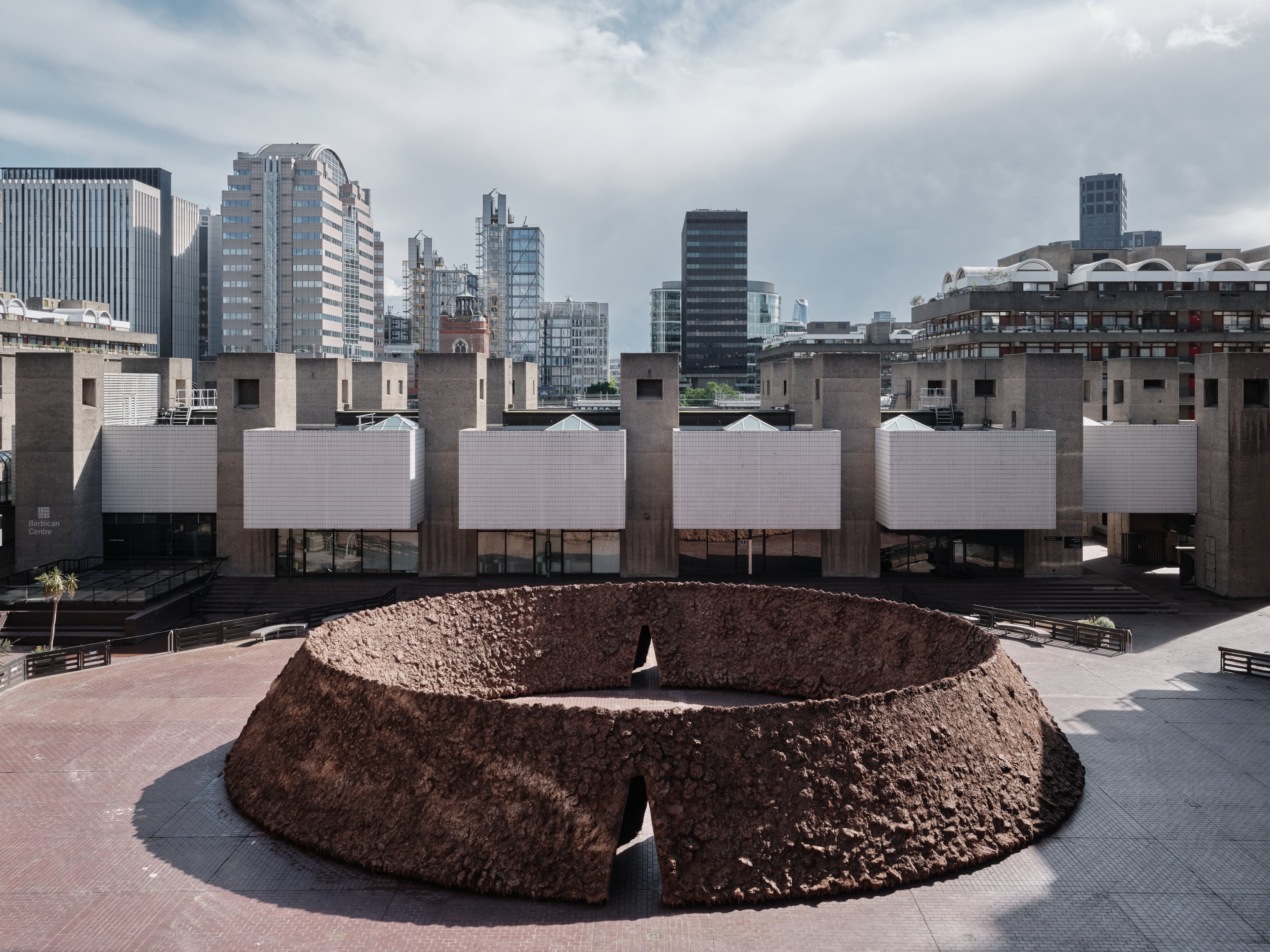 origo by Delcy Morelos, photographed by Thomas Adank — Elevated perspective showing the complete ovular ring with multiple entrances, London's glass towers visible beyond the brutalist complex.