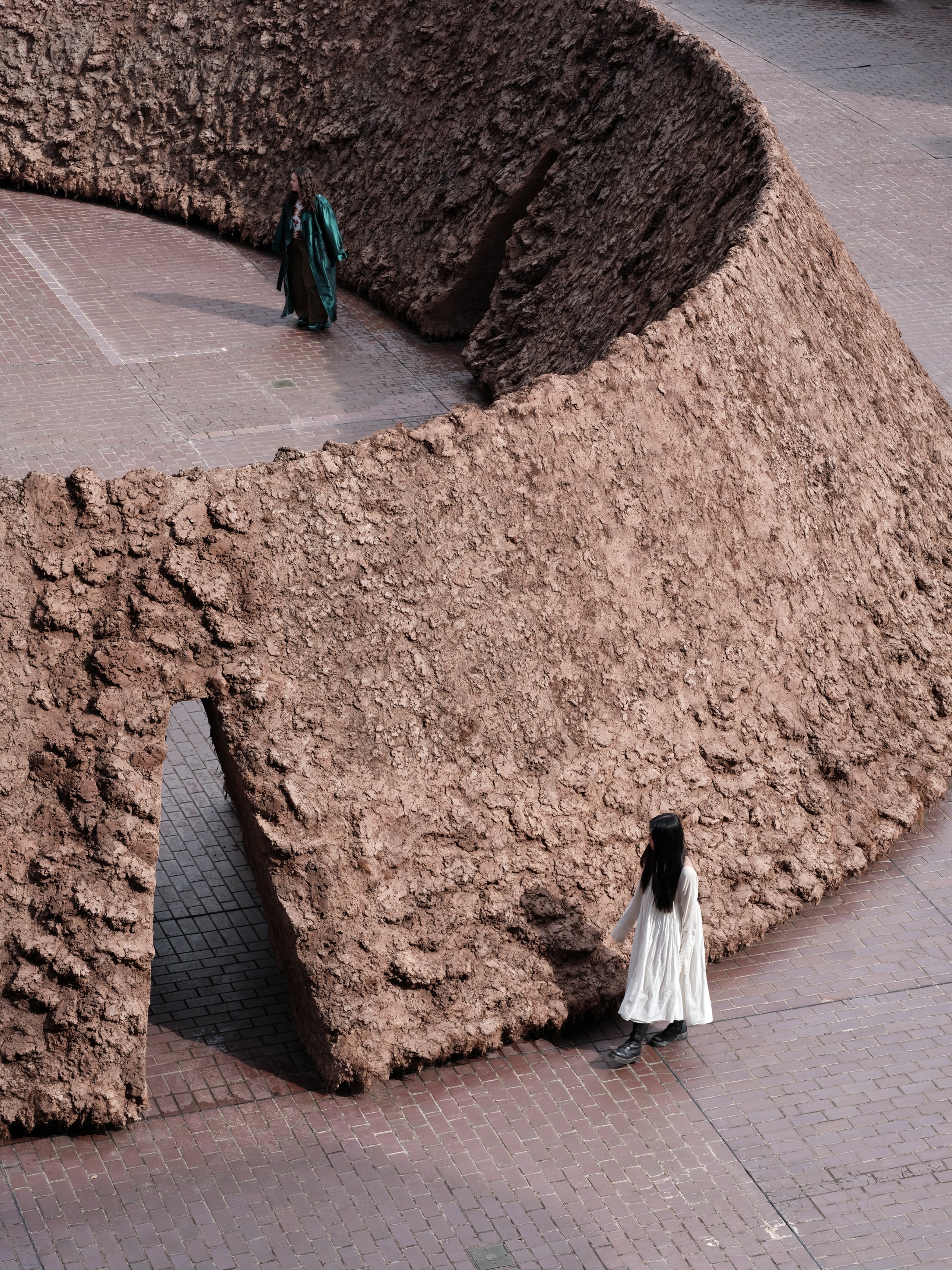 origo by Delcy Morelos, photographed by Thomas Adank — Aerial view of the curved pavilion with a figure in white entering through a triangular opening, hand-worked earth walls sweeping against brick paving.
