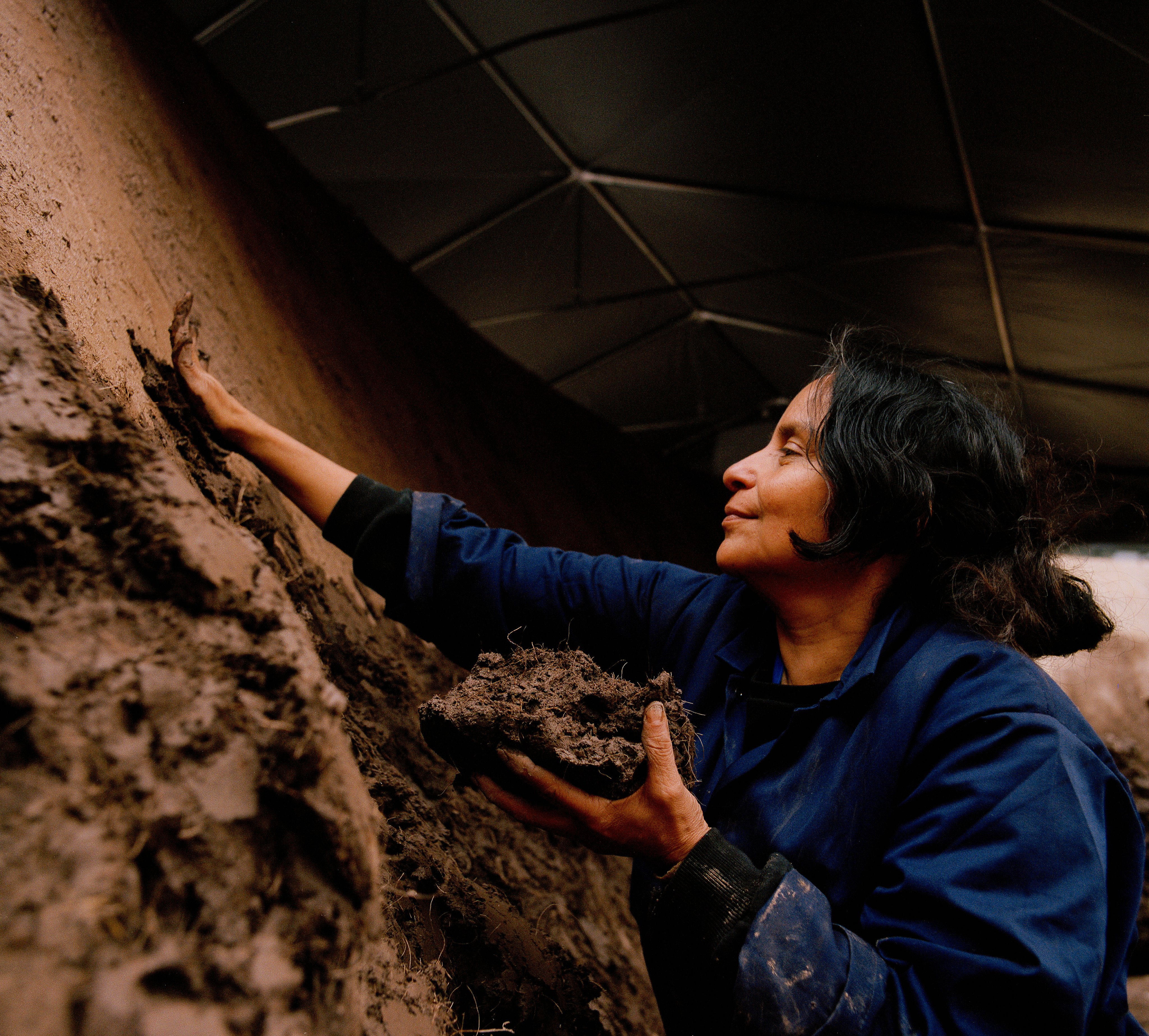 origo by Delcy Morelos, photographed by Thomas Adank — Close-up of Morelos' hands working clay mixed with hay fibers into the surface, face turned upward in concentration.