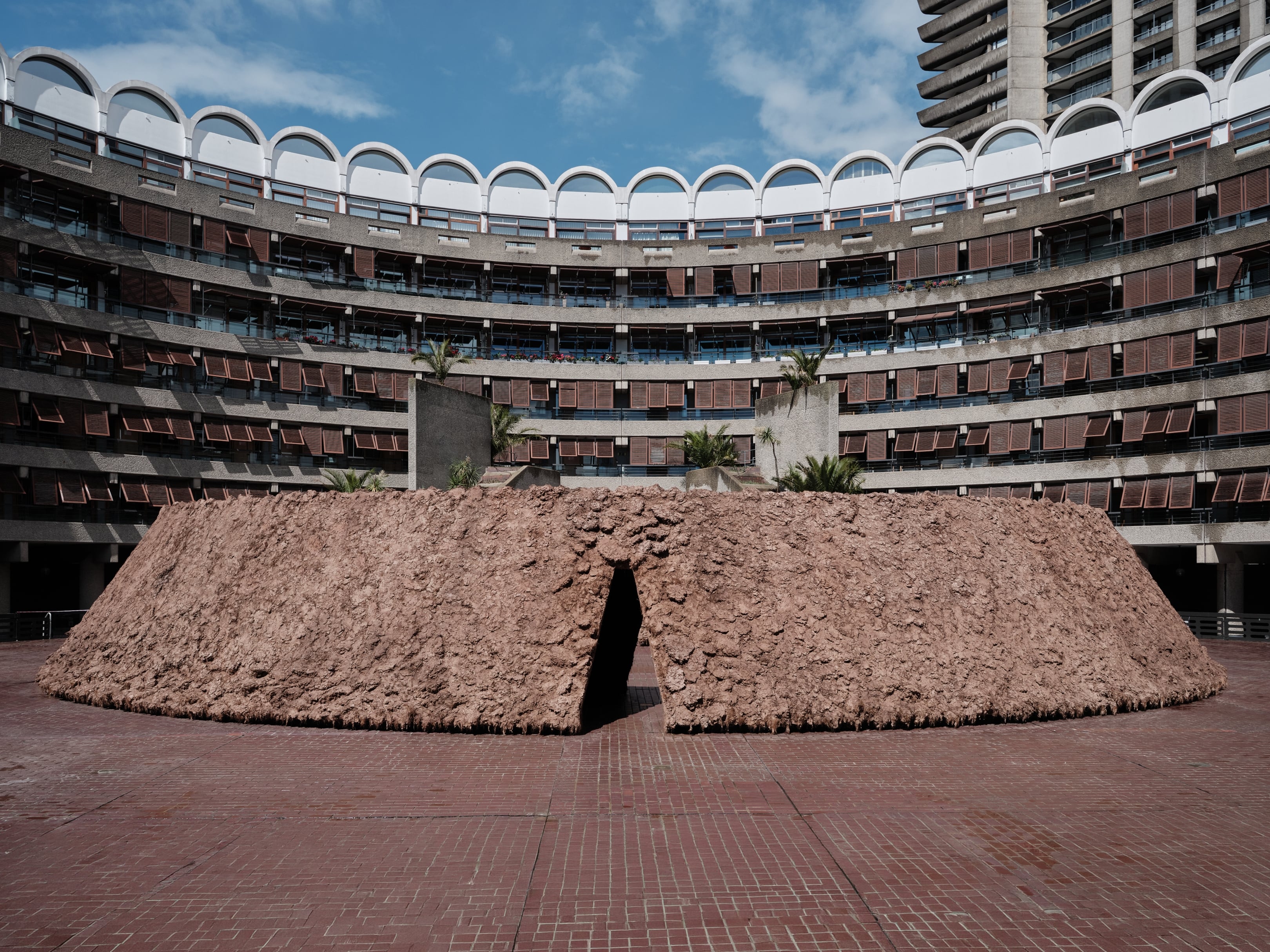 origo by Delcy Morelos, photographed by Thomas Adank — Full frontal view of the pavilion framed by the semicircular residential towers, brown shutters and concrete balconies encircling the earth form.