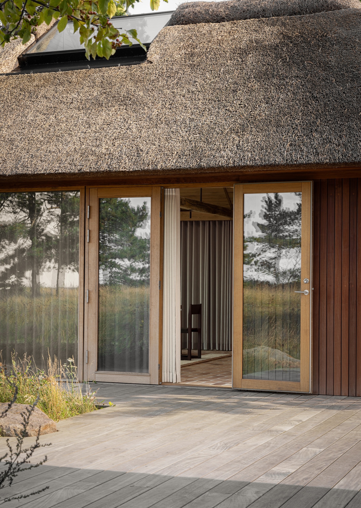 Shoreline Studio by Norm Architects, photographed by Norm Architects — Exterior entrance with thatched roof and skylight, vertical hardwood cladding, oak-framed glass doors, timber deck reflecting surrounding pines.
