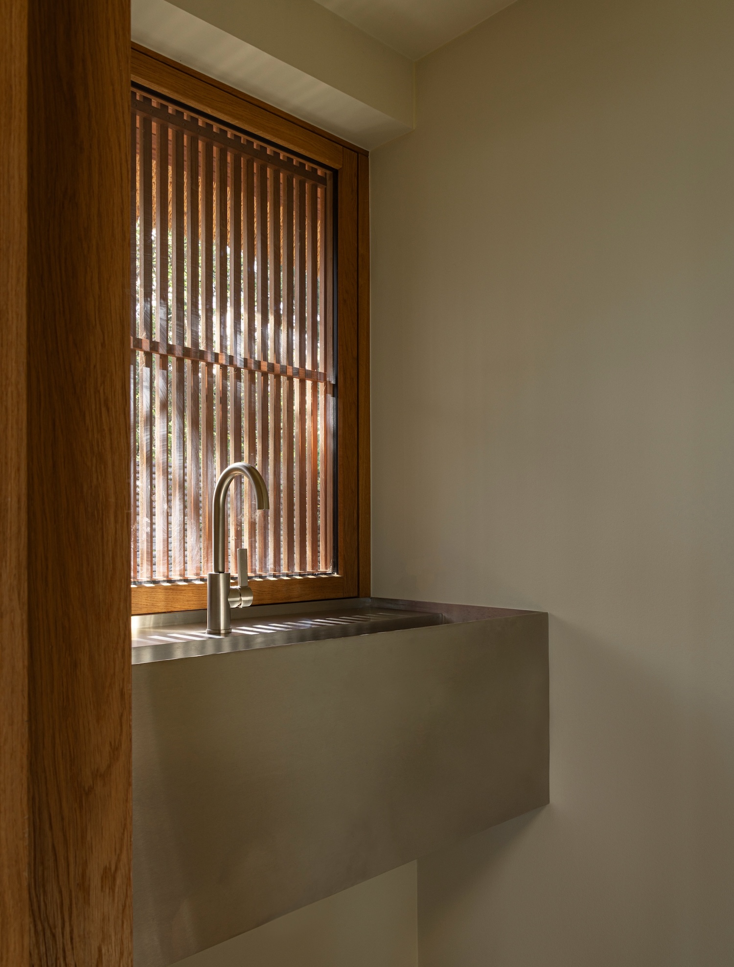 Shoreline Studio by Norm Architects, photographed by Norm Architects — Utility niche with stainless steel sink basin, oak-framed window with vertical wooden louvers casting striped shadows across the counter.