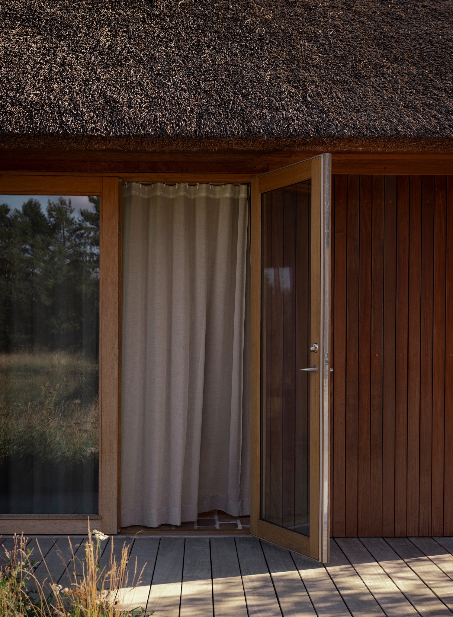 Shoreline Studio by Norm Architects, photographed by Norm Architects — Entrance threshold with thatched roof texture overhead, vertical hardwood cladding in warm brown, linen curtain visible through open glass door.