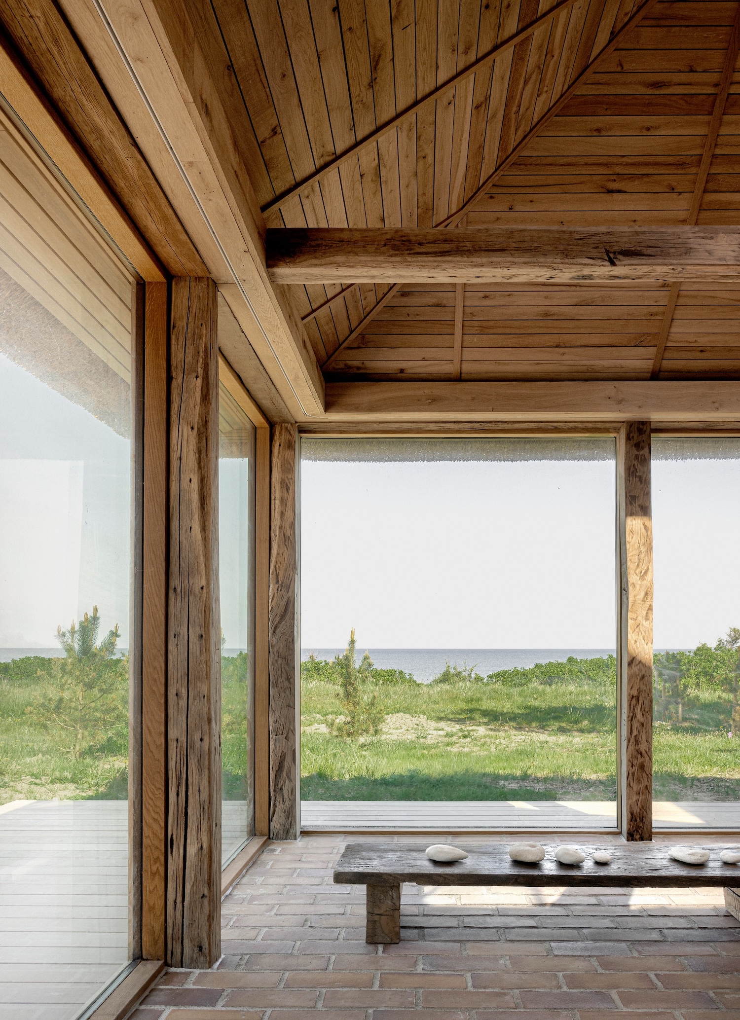 Shoreline Studio by Norm Architects, photographed by Norm Architects — Corner glazing framed by massive reclaimed timber posts revealing coastal landscape, brick floor and oak ceiling meeting at structural corner.