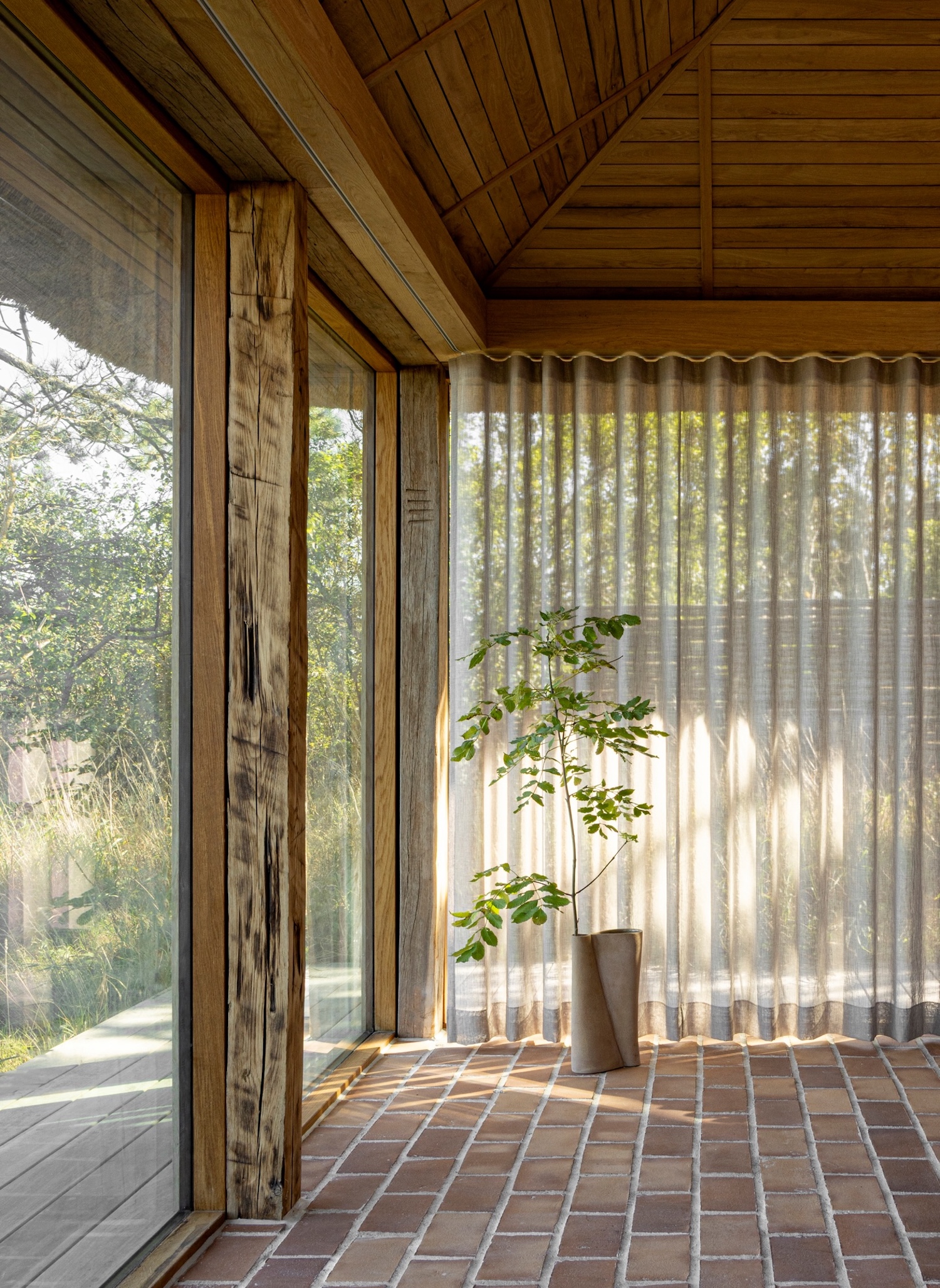 Shoreline Studio by Norm Architects, photographed by Norm Architects — Interior corner with reclaimed timber post and sheer curtains backlit by afternoon sun, potted plant in ceramic vessel on brick floor.