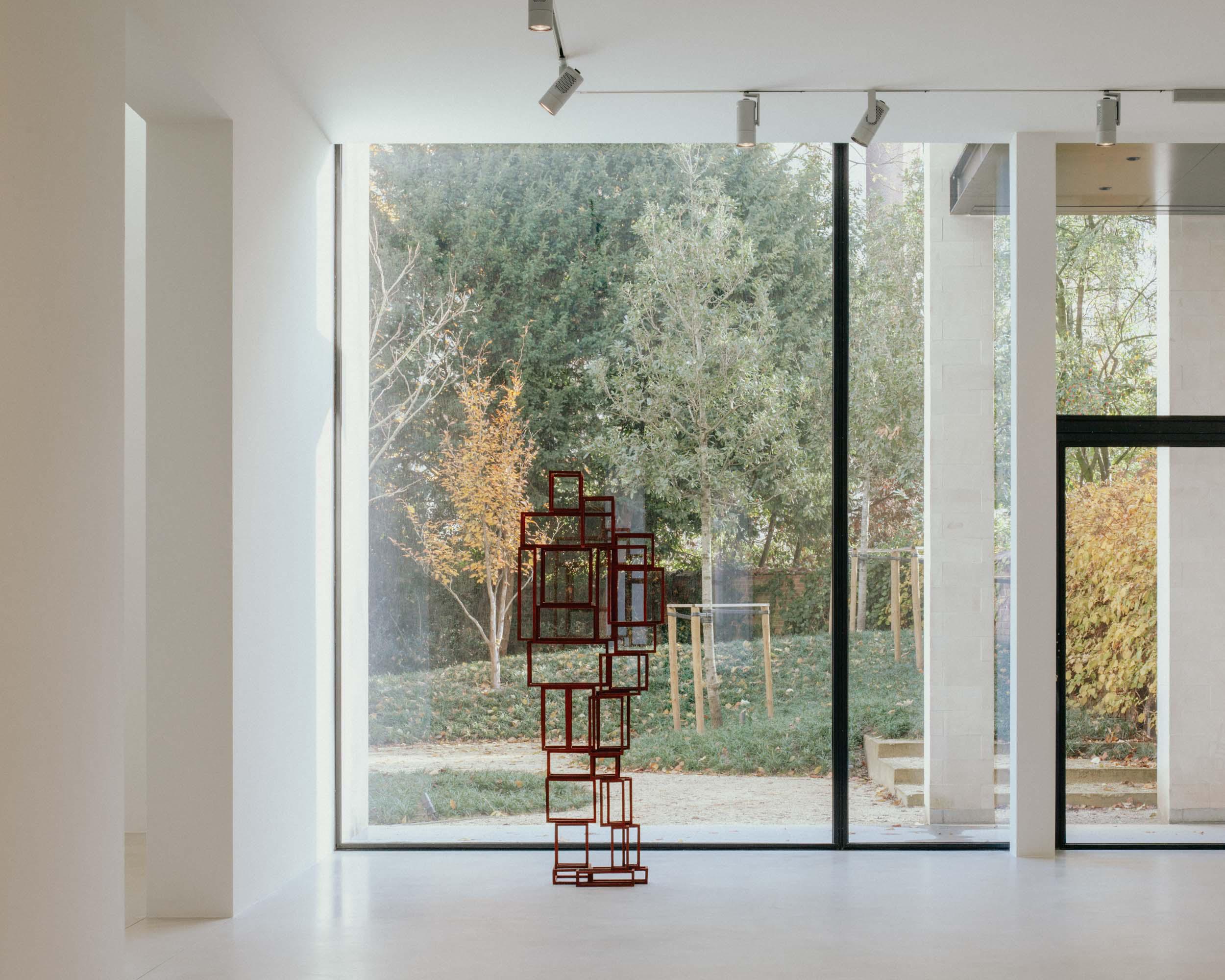Xavier Hufkens Gallery by Robbrecht en Daem Architecten, photographed by Jeremy Piret — Ground floor gallery with red steel frame sculptures silhouetted against floor-to-ceiling glass, garden visible, white polished concrete floor.