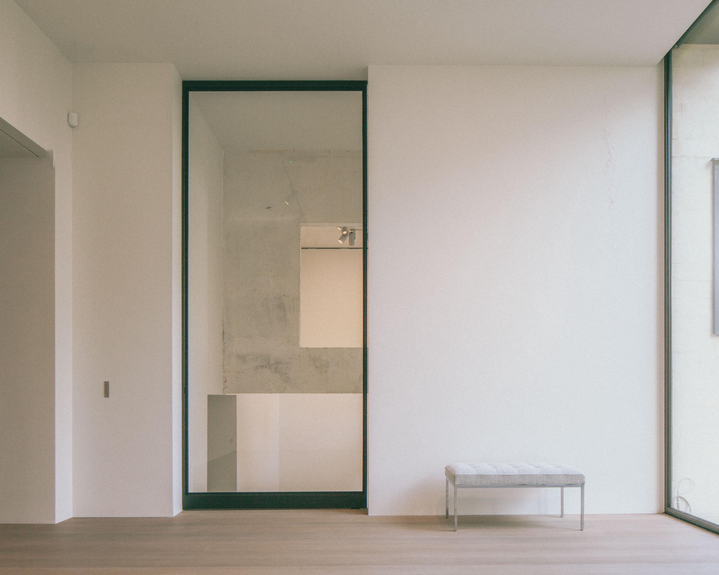 Xavier Hufkens Gallery by Robbrecht en Daem Architecten, photographed by Jeremy Piret — Gallery room with oak floor and dark-framed glazed door revealing concrete stairwell beyond, upholstered bench, side window.