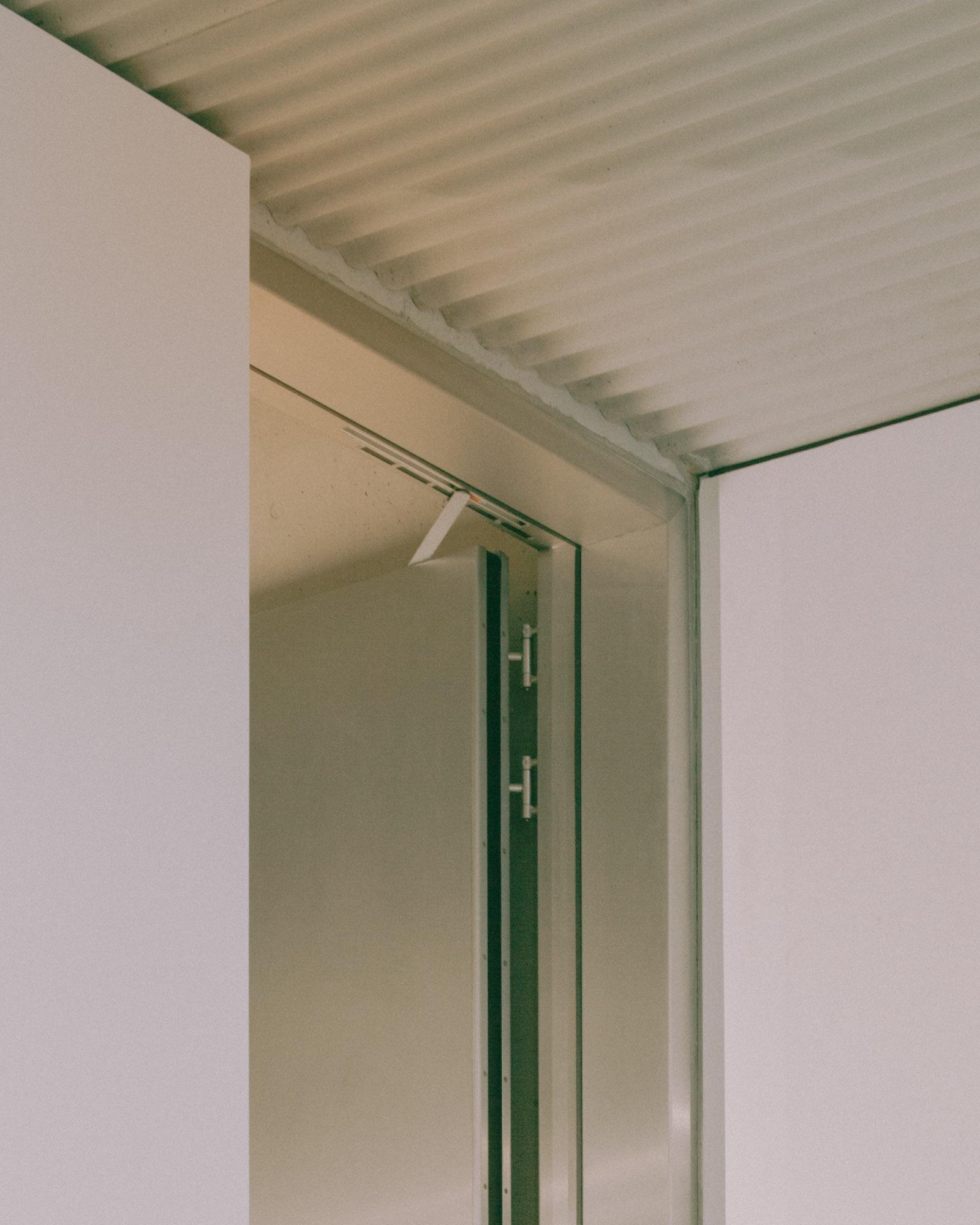 Xavier Hufkens Gallery by Robbrecht en Daem Architecten, photographed by Jeremy Piret — Junction of corrugated aluminum ceiling and white wall, dark green steel door frame, warm side light from gallery.