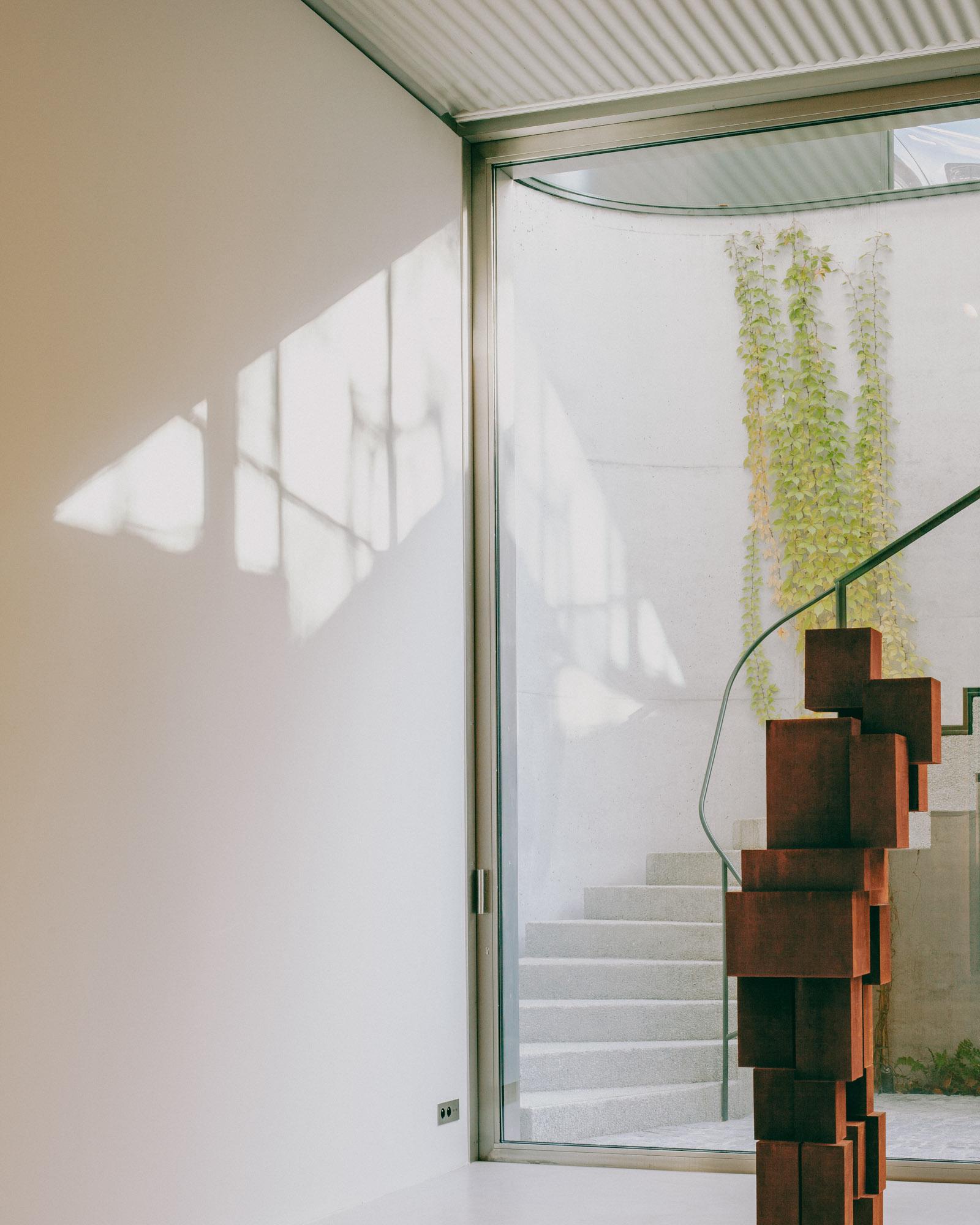 Xavier Hufkens Gallery by Robbrecht en Daem Architecten, photographed by Jeremy Piret — Interior looking through glass to external concrete stair with curved glass balustrade, corten sculpture, trailing vines on concrete walls.