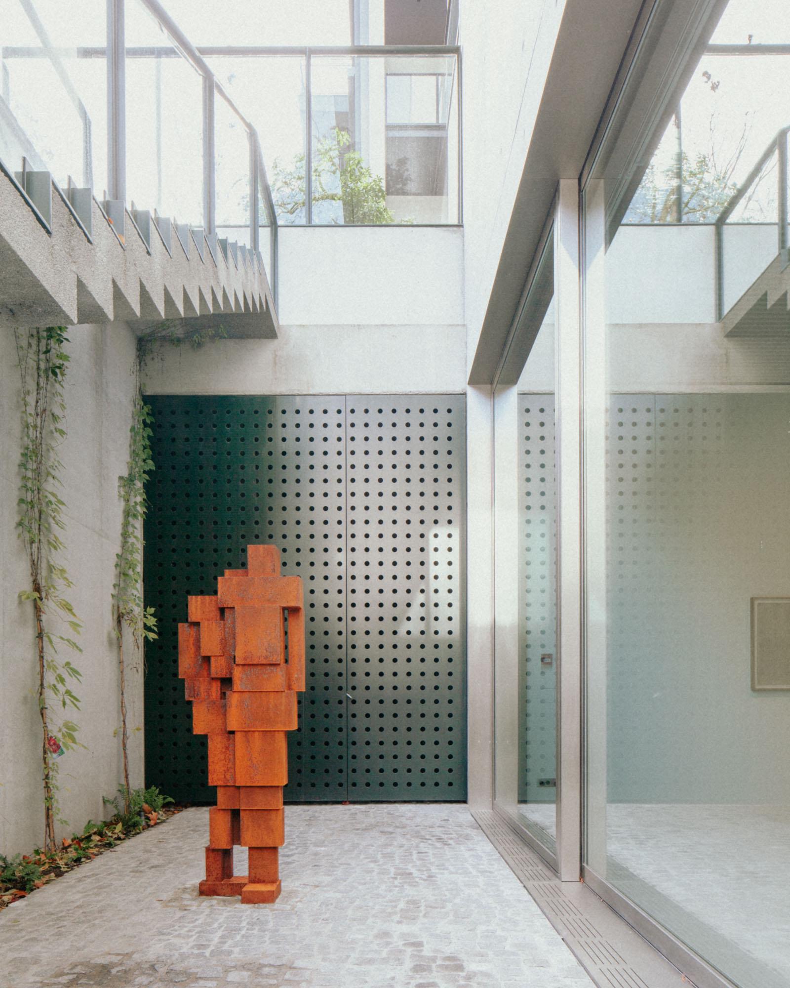 Xavier Hufkens Gallery by Robbrecht en Daem Architecten, photographed by Jeremy Piret — Courtyard with corten figure sculpture against perforated dark green metal screen, Belgian cobblestones, concrete walls, trailing vines above.