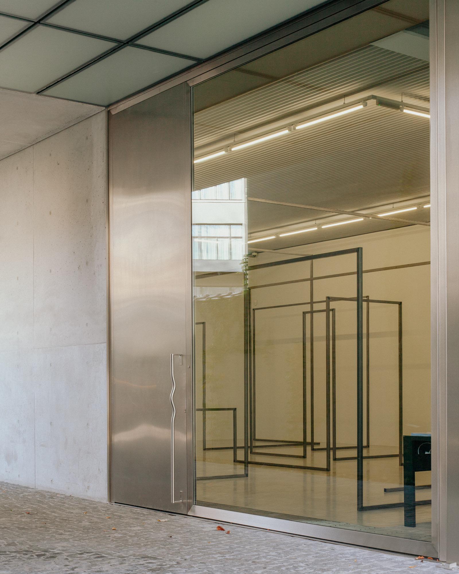Xavier Hufkens Gallery by Robbrecht en Daem Architecten, photographed by Jeremy Piret — Entrance detail with stainless steel door and glass beside fair-faced concrete, linear steel frame sculptures visible inside corrugated-ceiling gallery.