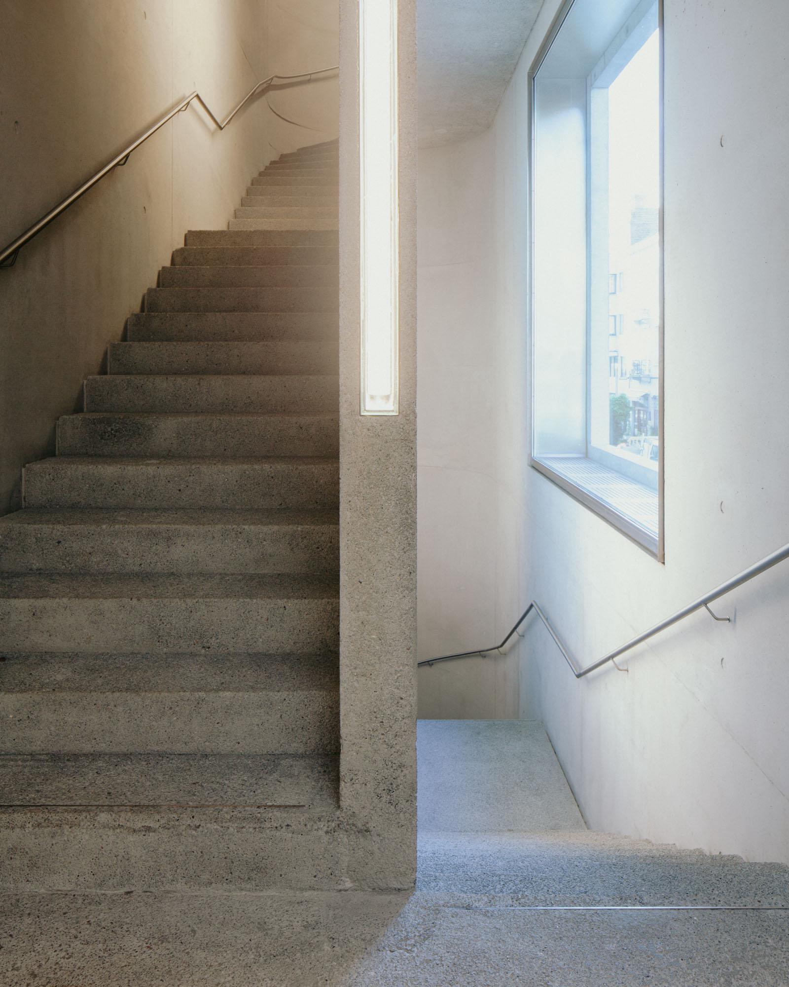 Xavier Hufkens Gallery by Robbrecht en Daem Architecten, photographed by Jeremy Piret — Split view of two concrete stair runs with aggregate finish, steel handrail, strip lighting, window washing the landing.