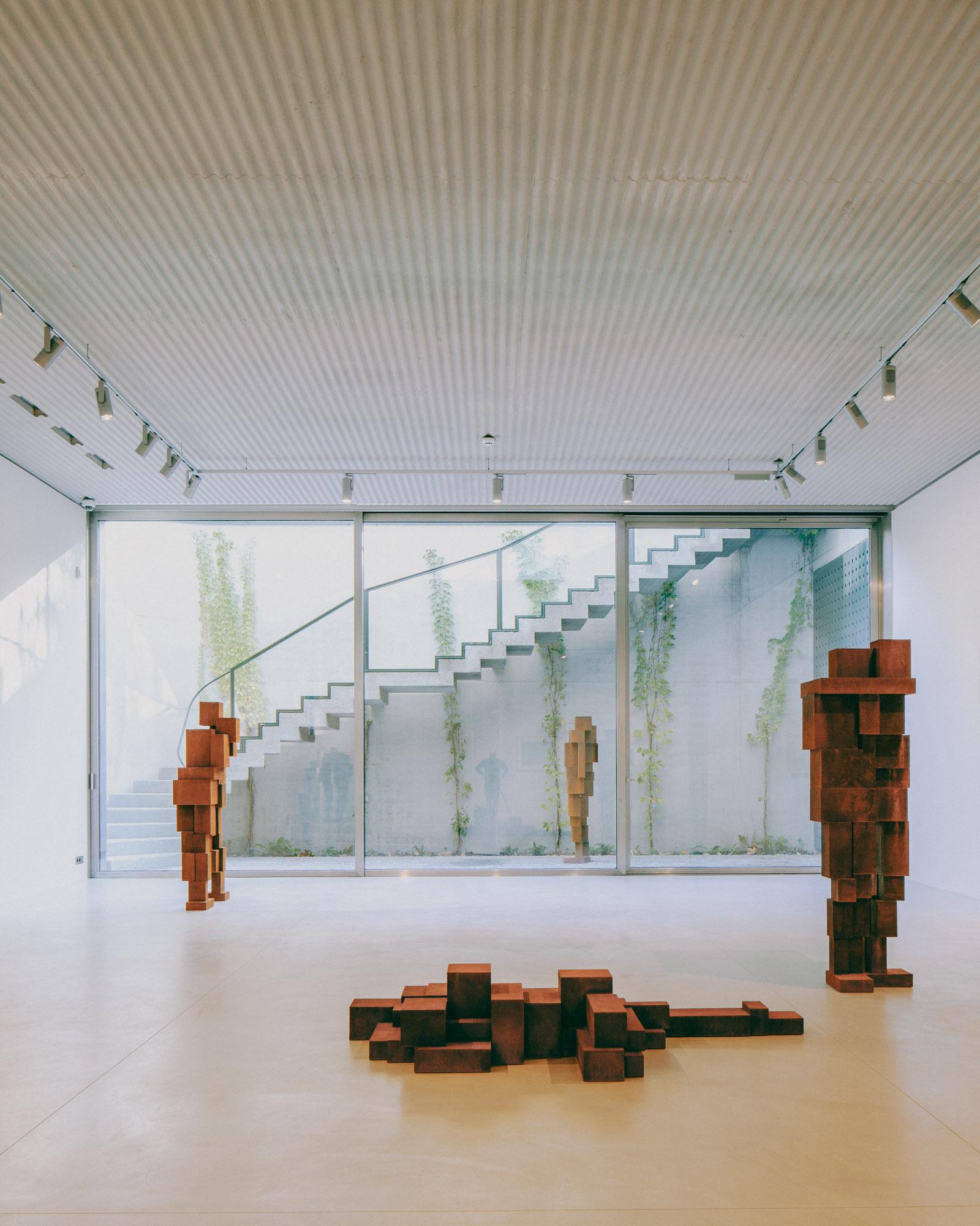 Xavier Hufkens Gallery by Robbrecht en Daem Architecten, photographed by Jeremy Piret — Main gallery with corten steel figure sculptures, corrugated aluminum ceiling, glass wall to courtyard where external stair rises behind trailing vines.