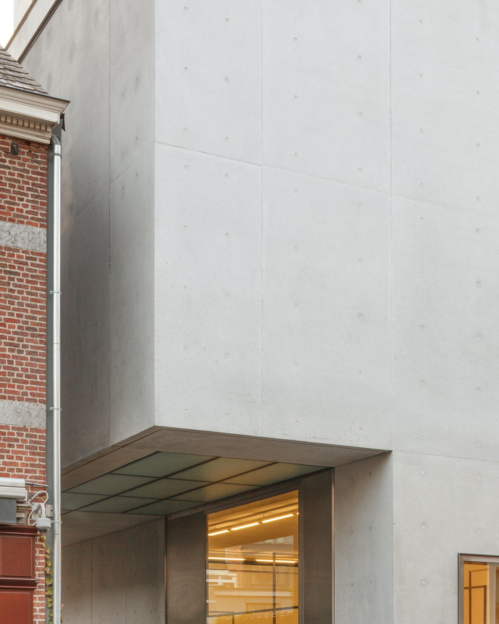 Xavier Hufkens Gallery by Robbrecht en Daem Architecten, photographed by Jeremy Piret — Entrance canopy with fair-faced concrete and visible tie holes, glass ceiling revealing warm interior light, brick neighbor beyond.